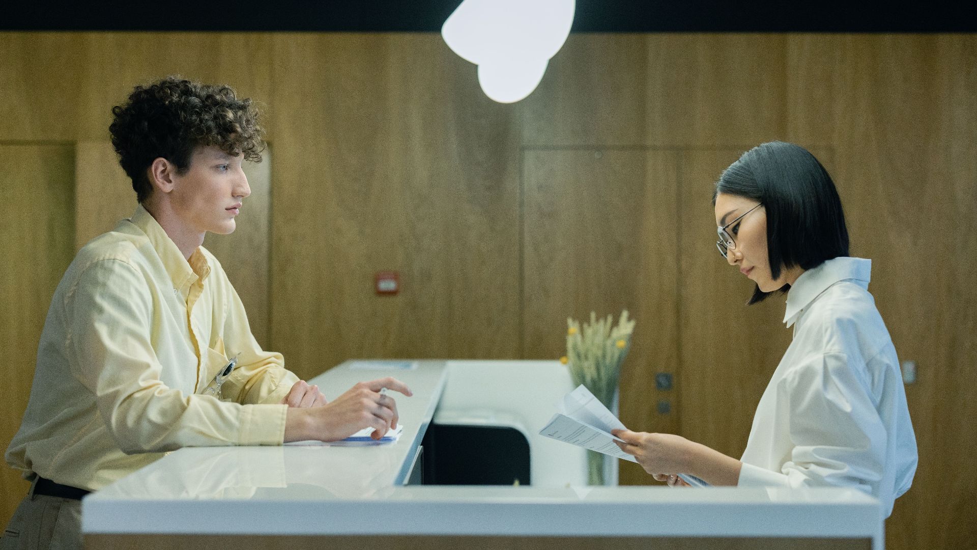 A young man and woman interacting at a modern office reception desk indoors.
