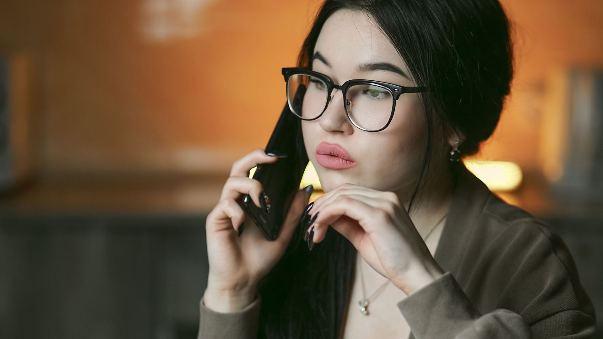 A woman with eyeglasses engaged in a phone call indoors, focused and thoughtful.