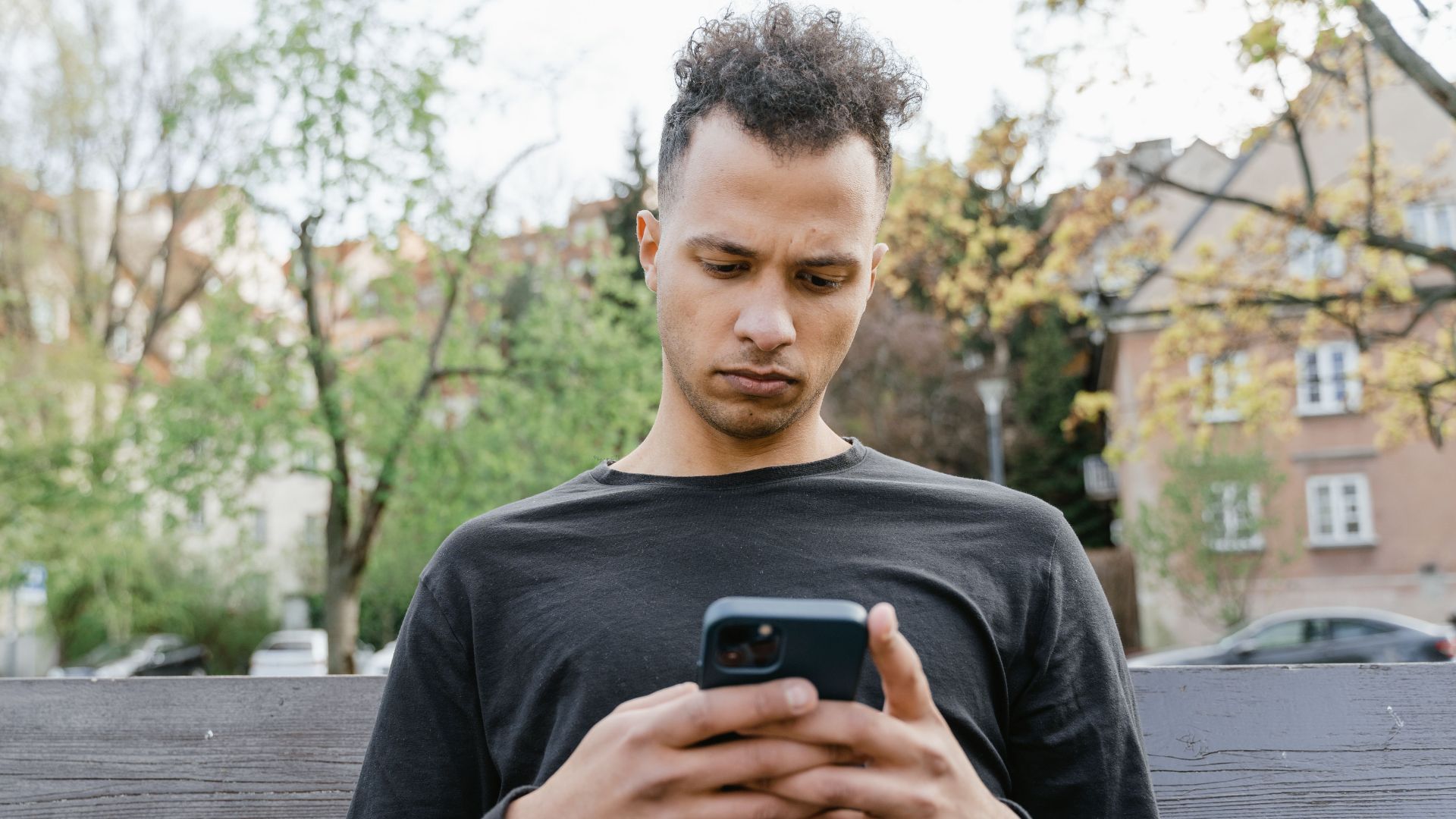 Casual young man sitting on a bench, texting on his smartphone in a park setting.
