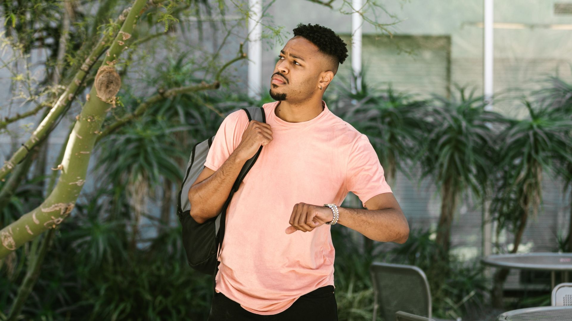 A young man in a casual outfit checks the time while walking outdoors with a backpack.