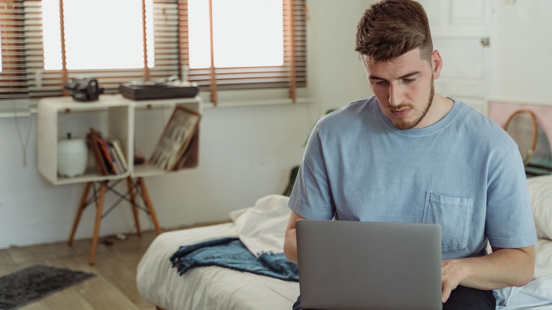 A bearded man in a blue t-shirt focused on a laptop in a sunlit bedroom.