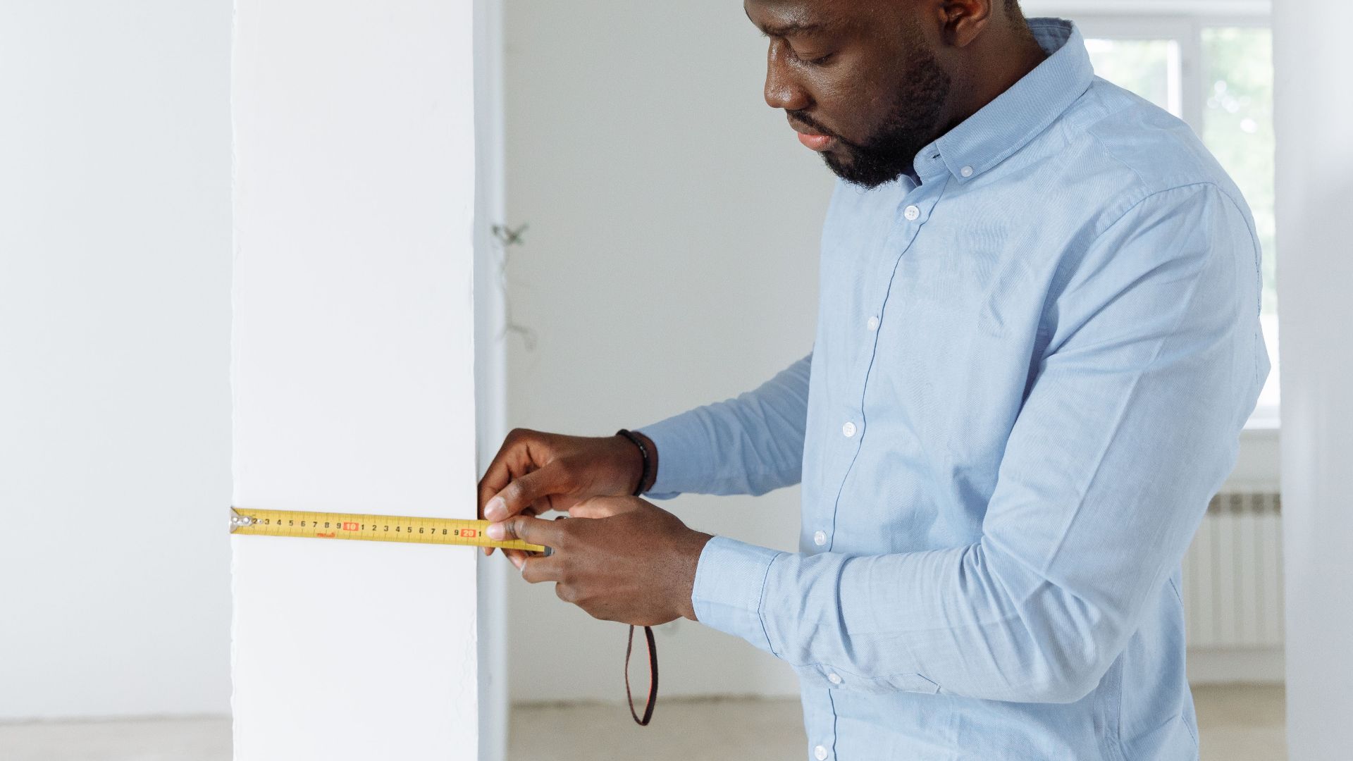 Construction worker measuring a wall indoors, wearing a white hard hat and blue shirt.