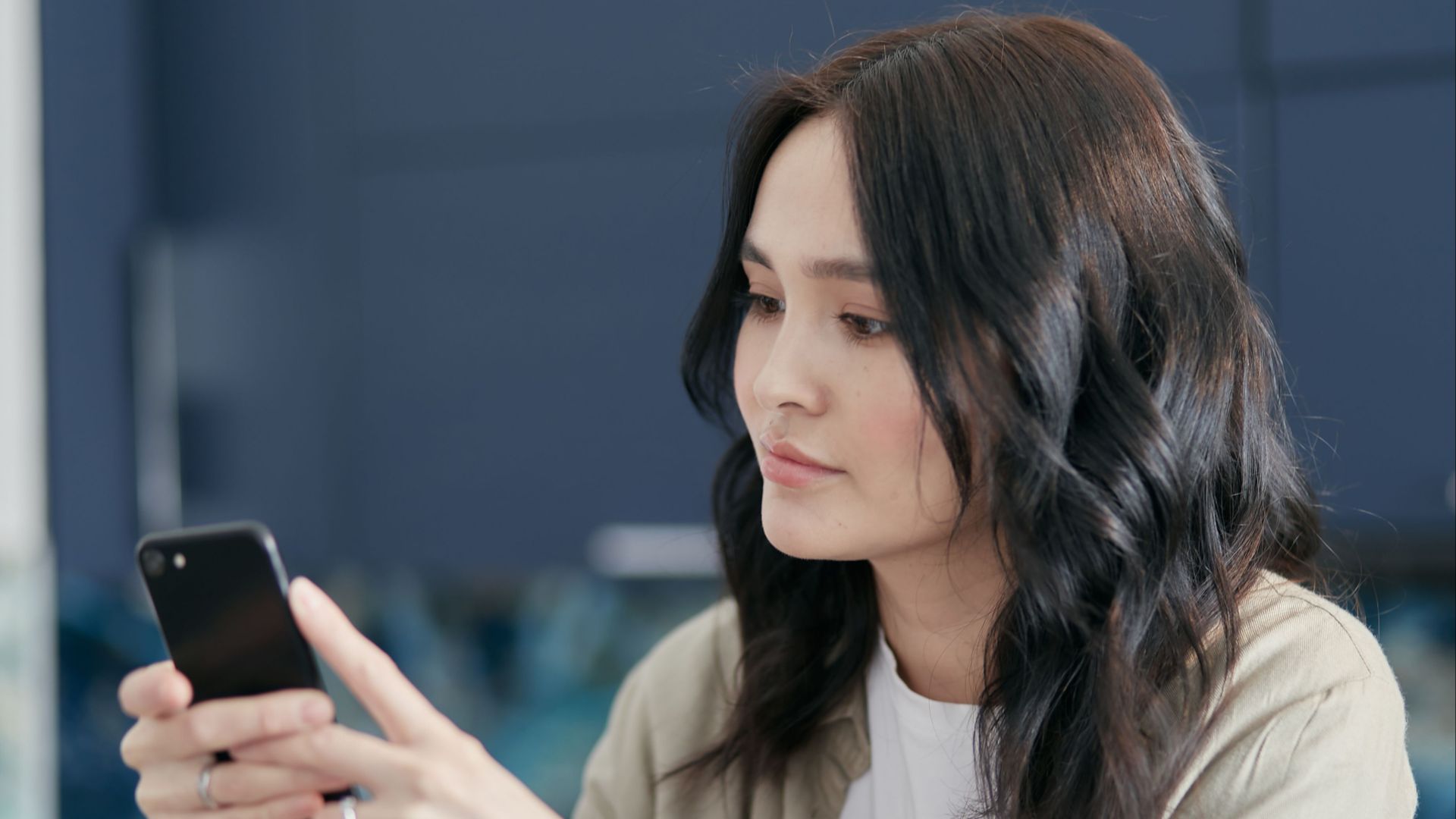 A young woman with black hair using a smartphone indoors, focused on the screen.