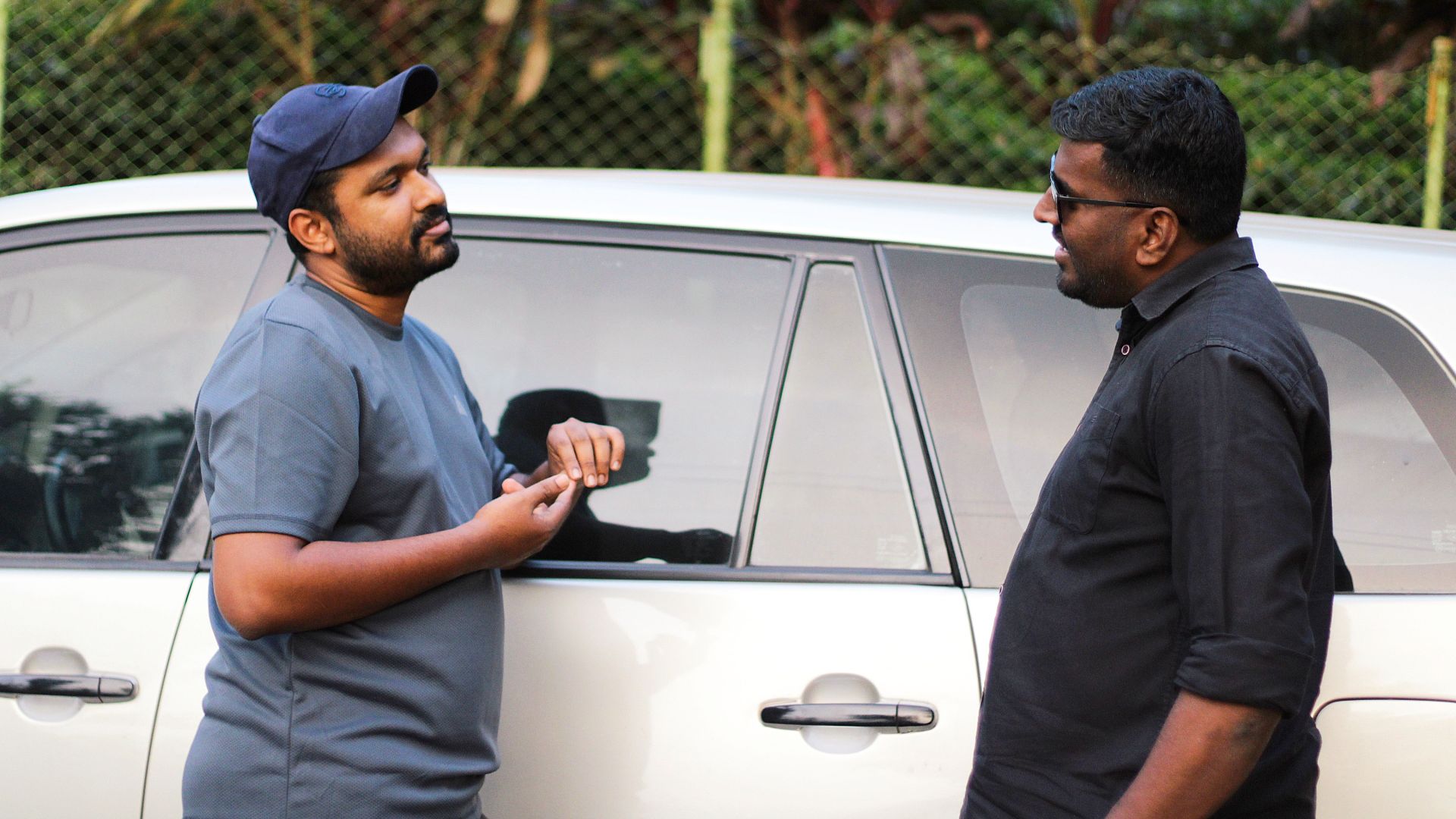Two men having a casual discussion beside a car in an outdoor setting.