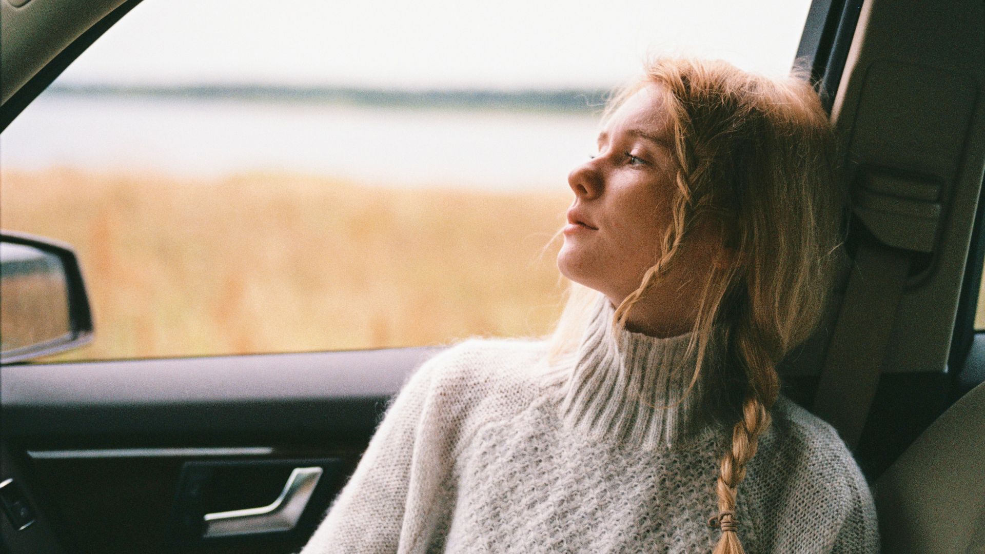 A thoughtful woman with braided hair gazes out a car window during a daytime road trip.