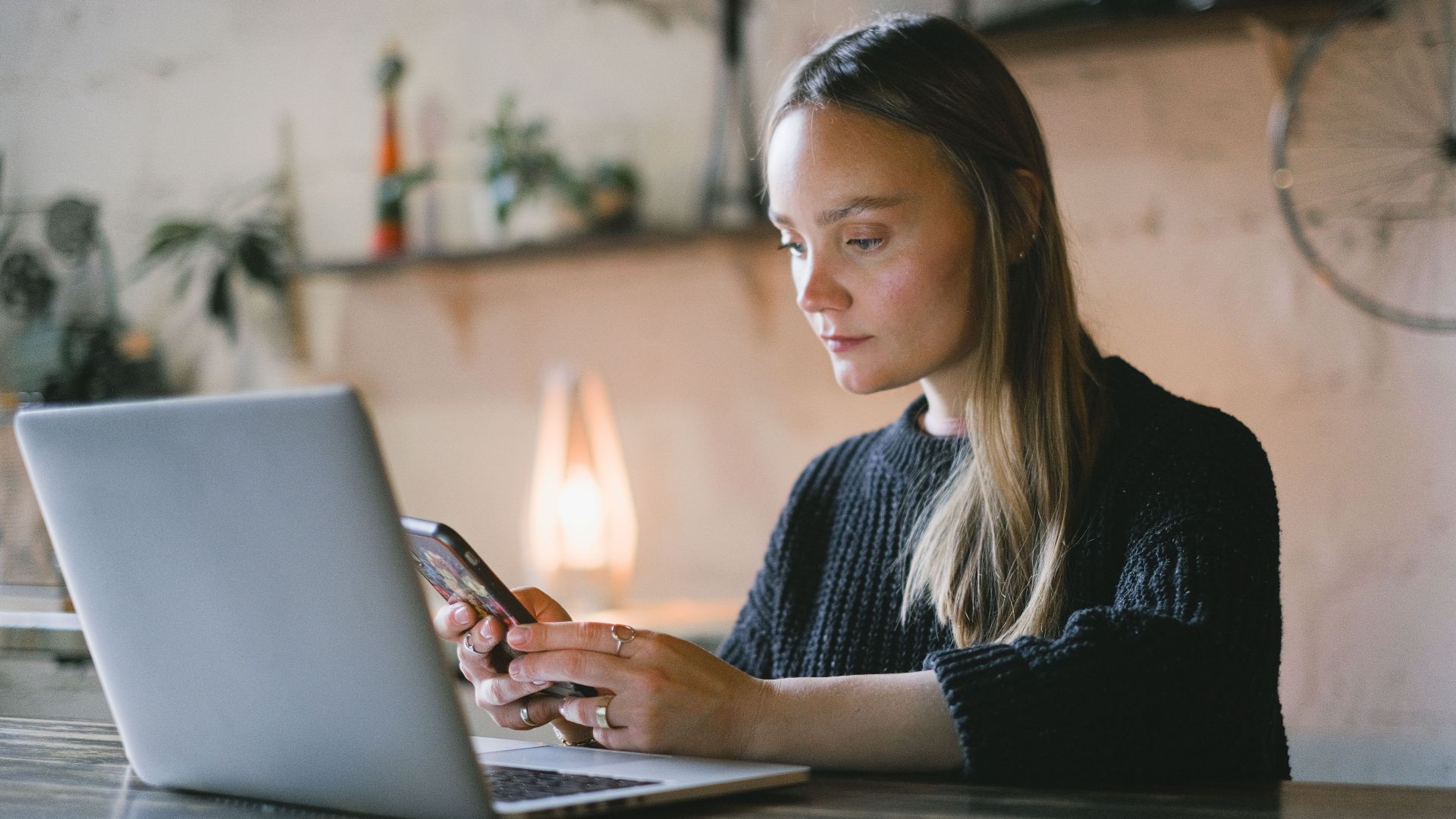Concentrated female freelancer text messaging on cellphone while sitting at table with netbook during online work on blurred background