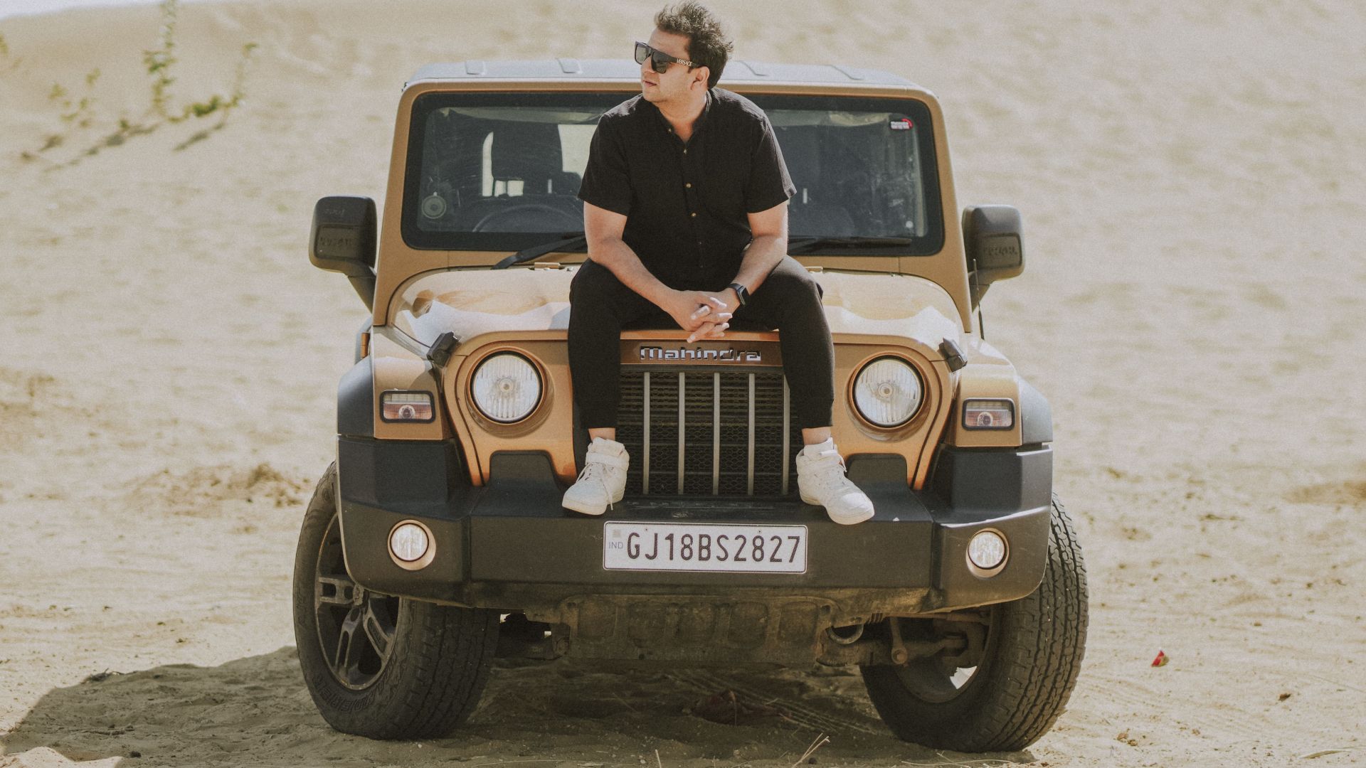 A man sits on a modern SUV parked in a sandy desert, wearing sunglasses and casual attire.