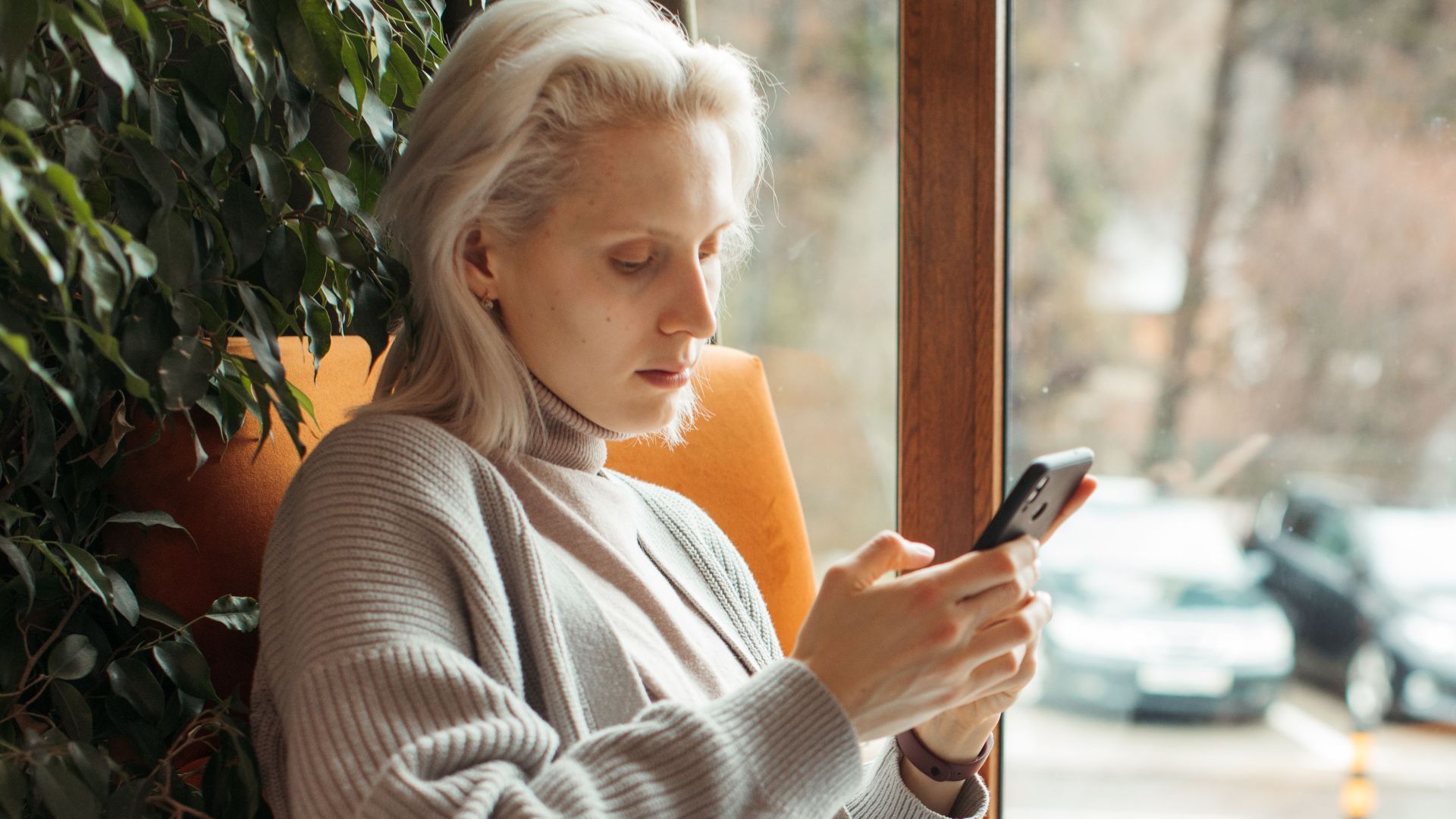 Woman sitting by a window, focused on her smartphone and laptop, in a cozy setting.