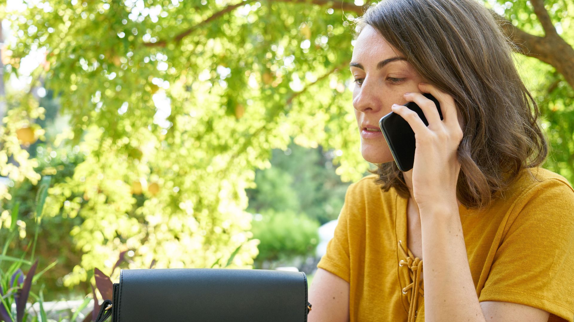 A woman in a yellow shirt makes a phone call outdoors amid lush greenery.