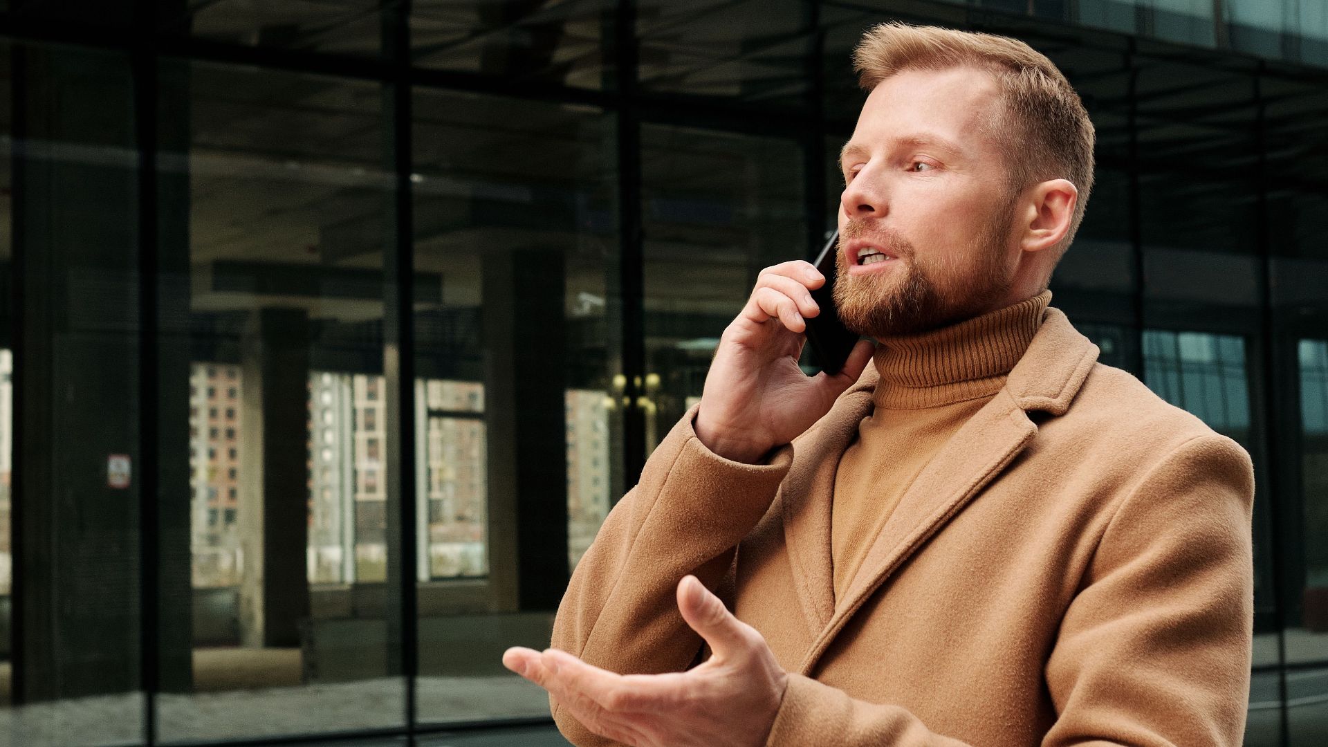 Confident businessman in brown coat speaking on phone outdoors against modern glass building.