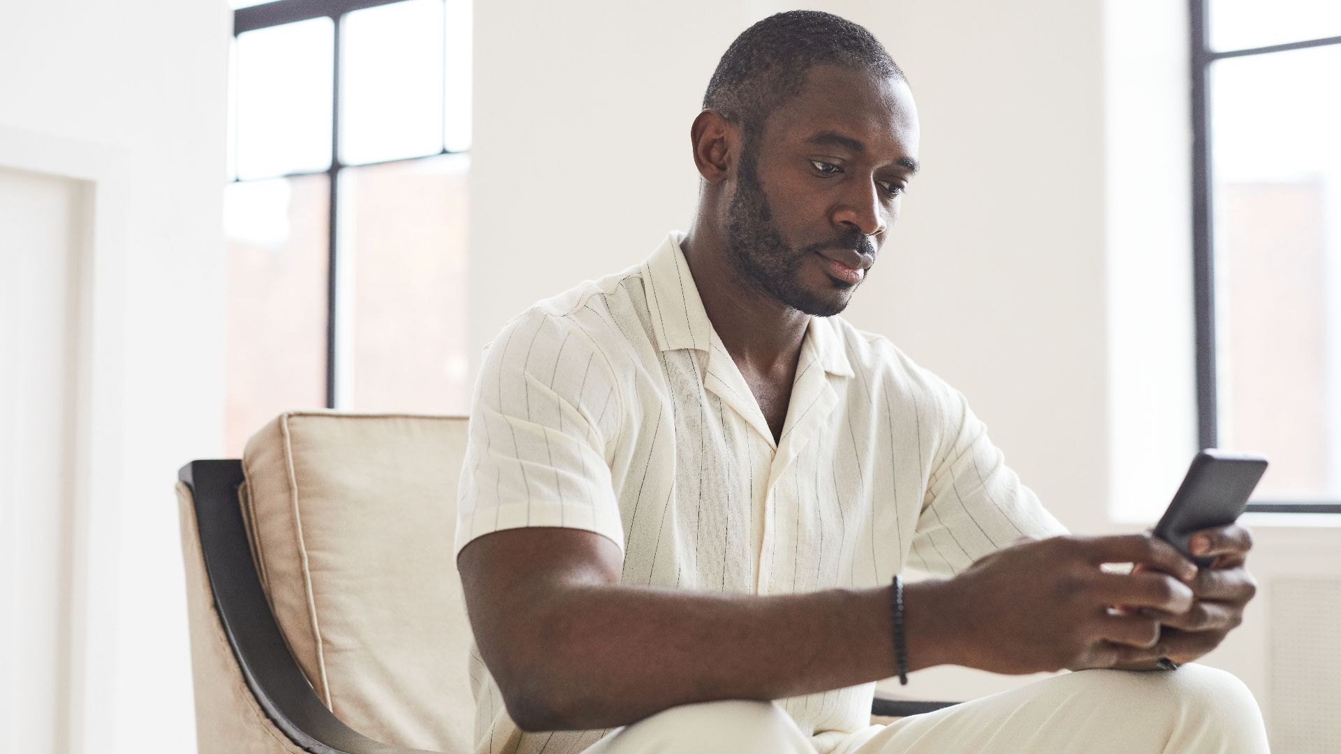 Man with facial hair sitting indoors using smartphone, focusing on screen.