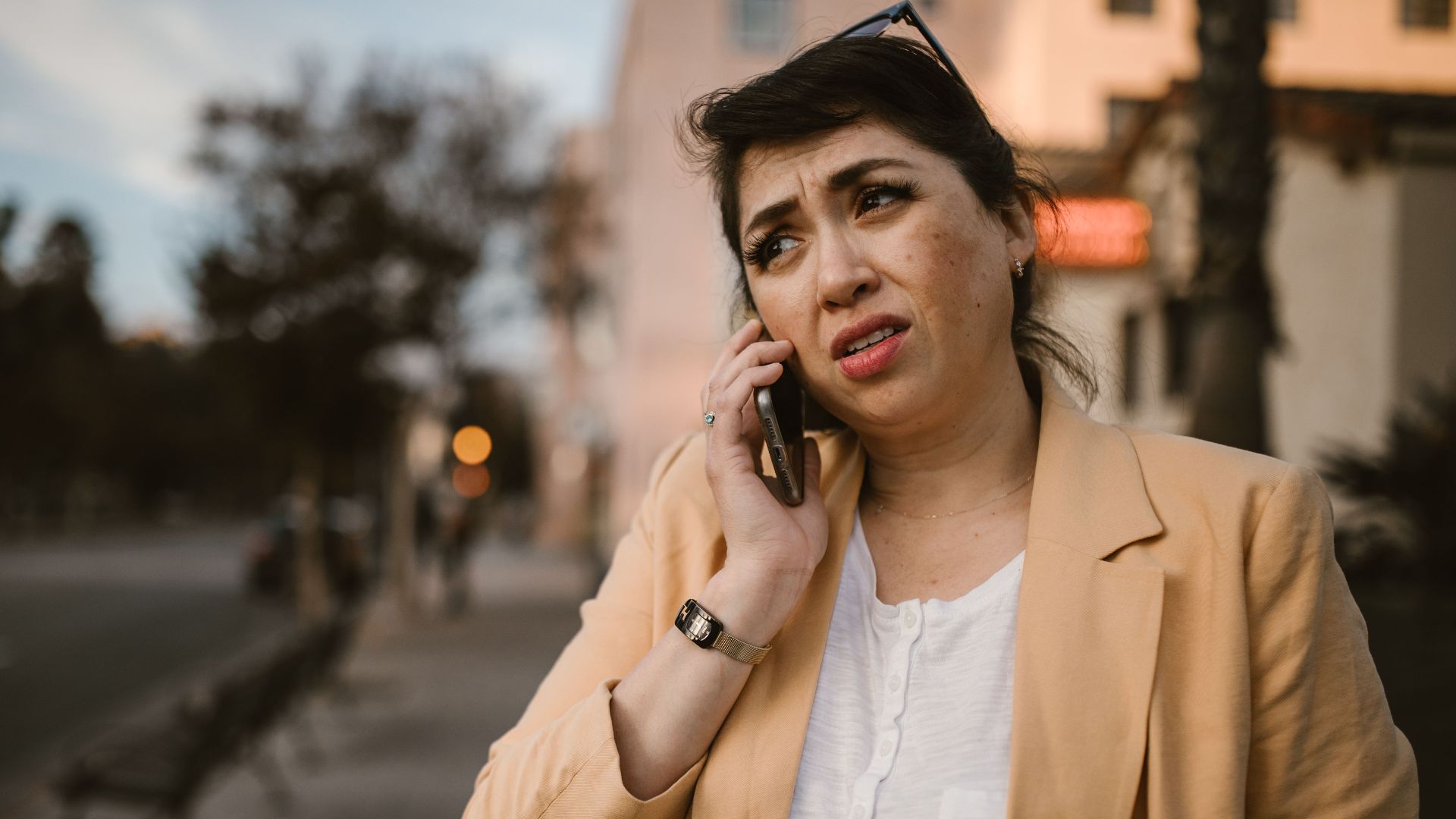 Woman in beige jacket looks concerned while making phone call outdoors.