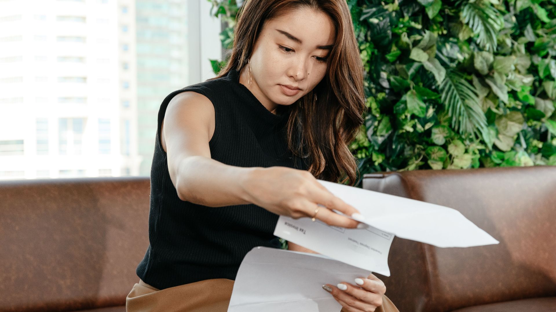 Focused Asian female turning pages of document while sitting on sofa during paperwork in modern workspace with green deciduous plant