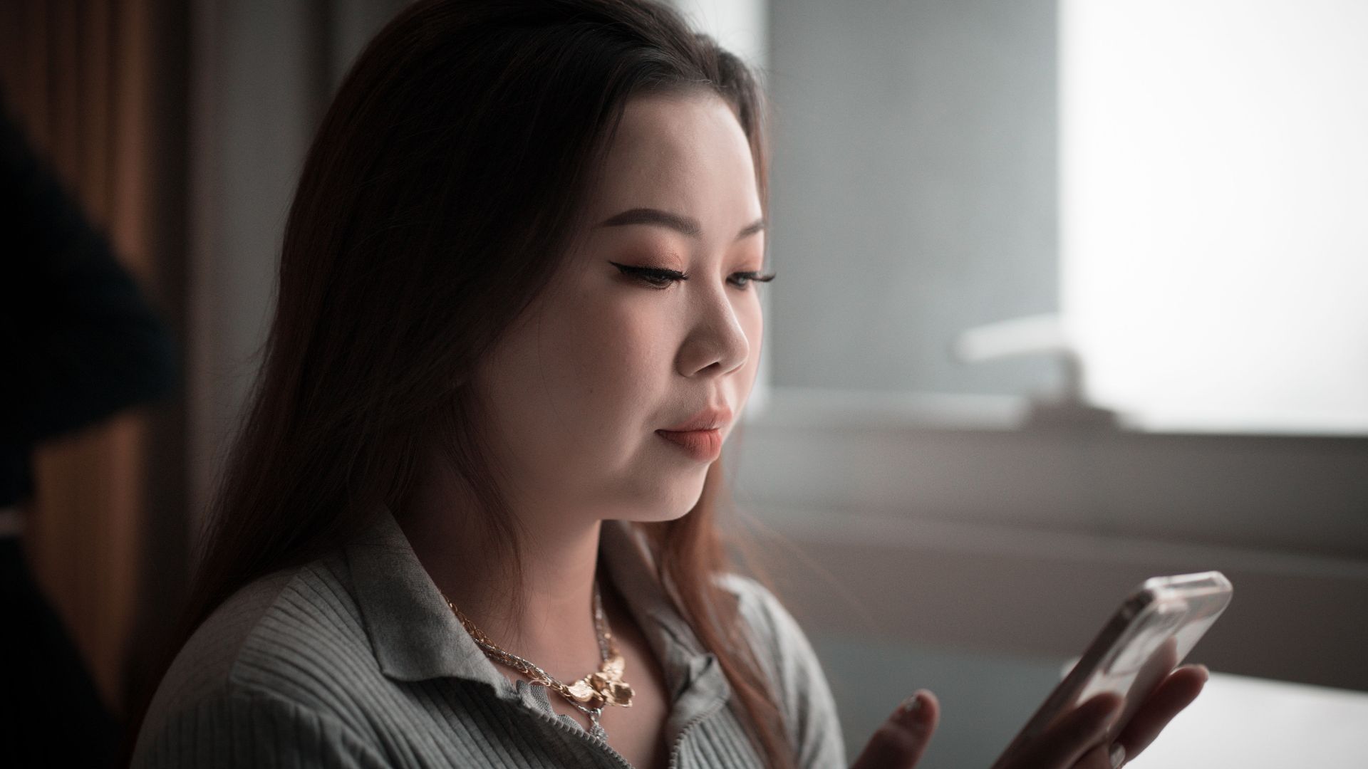 Young Asian woman using smartphone indoors, focused on screen.
