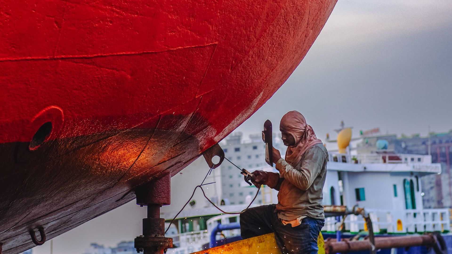 A worker repairs a large ship's hull at a bustling Dhaka shipyard, showcasing skilled craftsmanship.