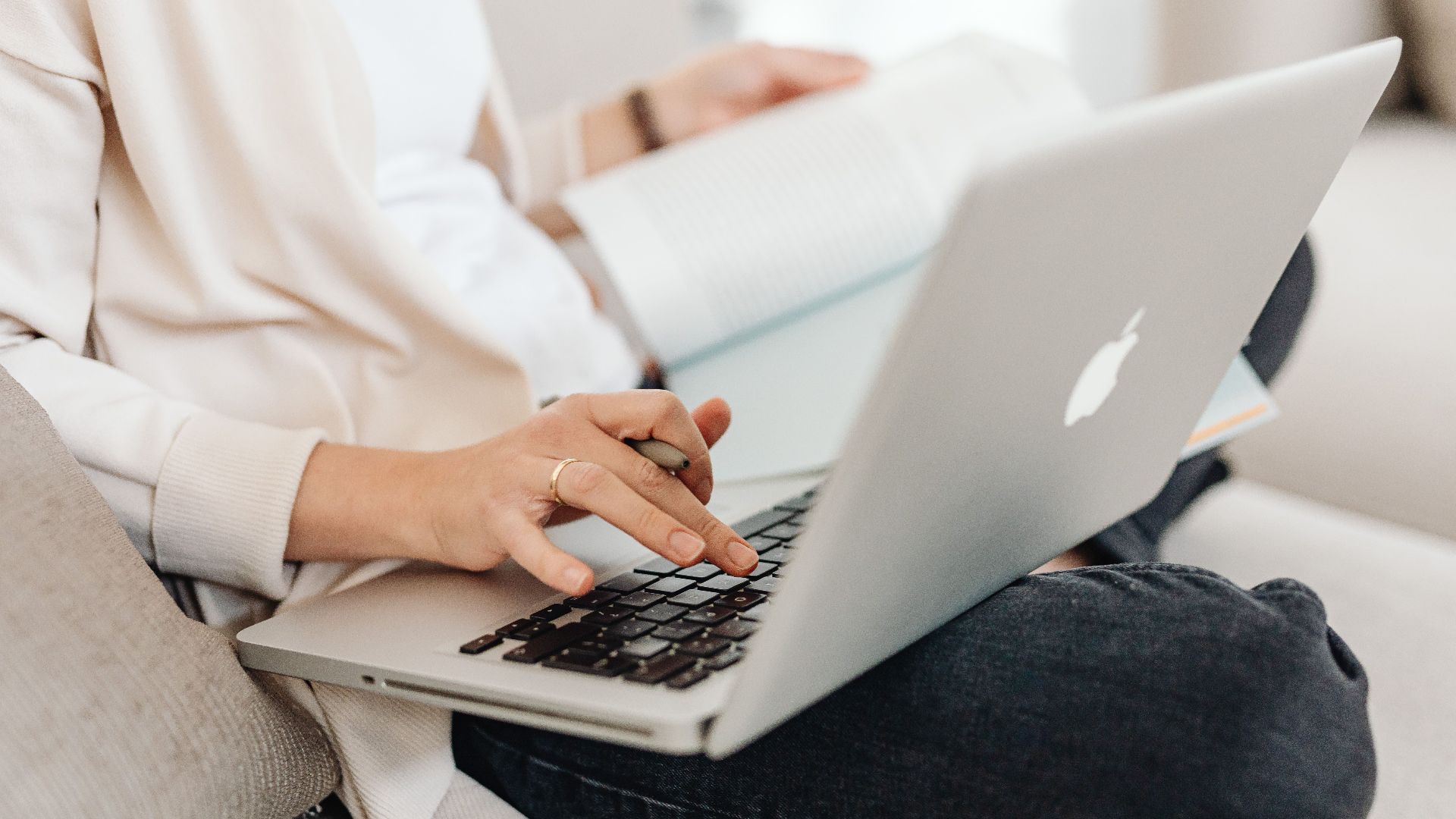 Close-up of a person multitasking, typing on a laptop while holding a book on their lap.