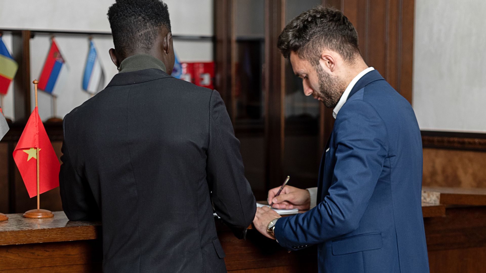 Two men in suits signing documents at a hotel reception desk, indicating partnership and professionalism.