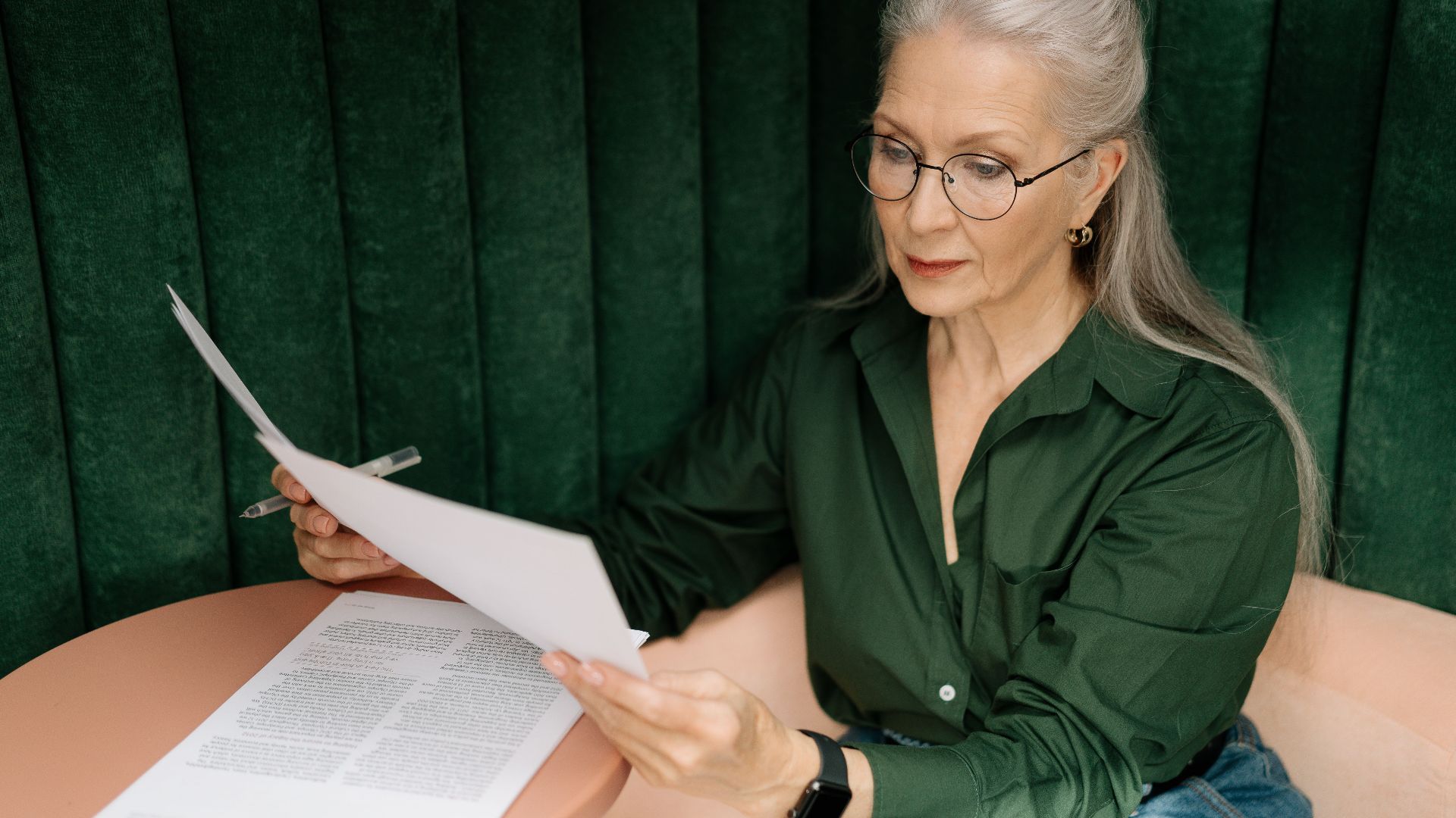 Senior woman wearing glasses, reading documents at a desk indoors, showcasing professional focus.