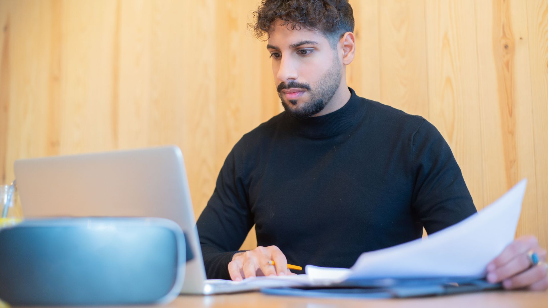Focused businessman with beard working on a laptop, reviewing documents in a modern workspace.
