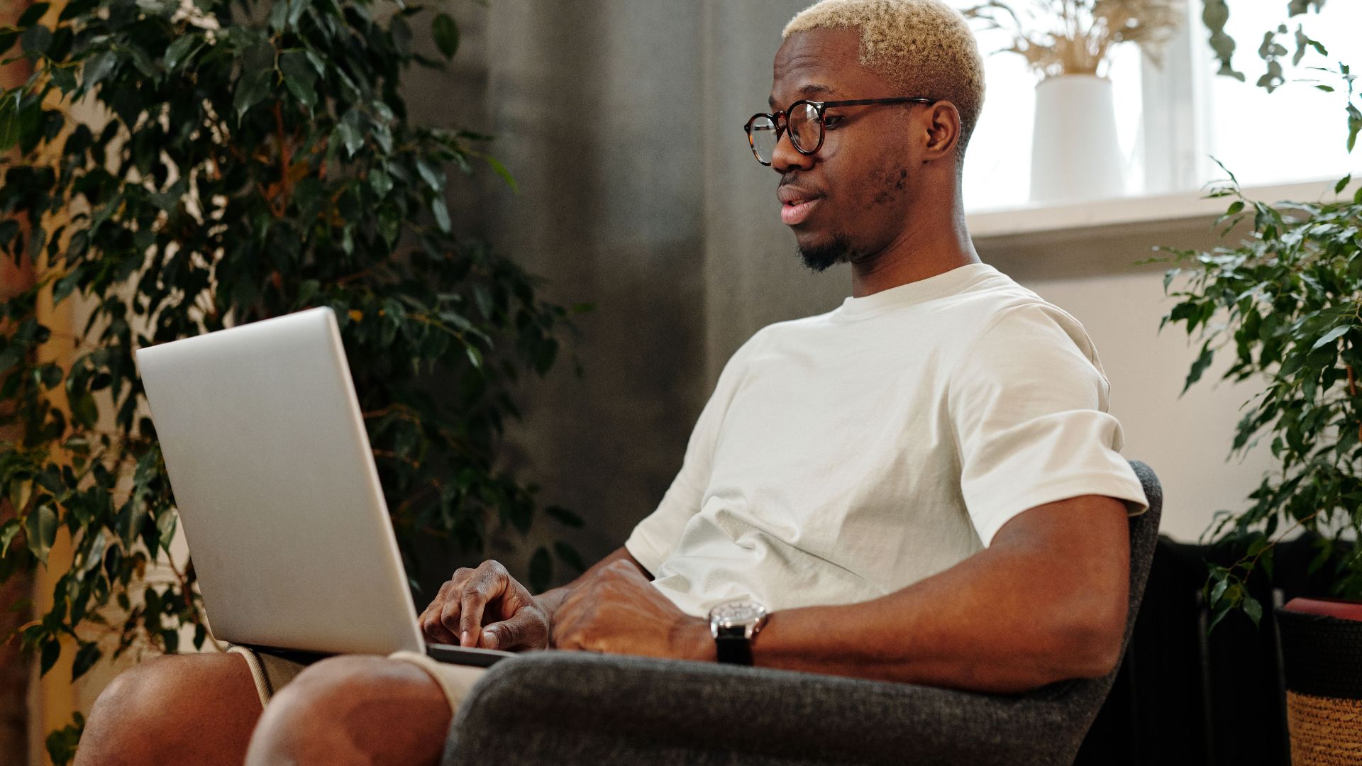 Focused young man with glasses working on a laptop in a cozy indoor space surrounded by plants.