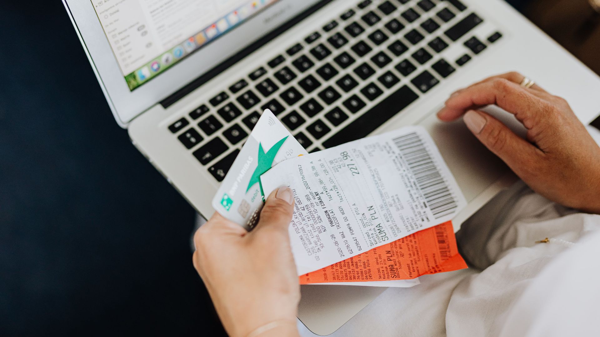 Close-up of hands holding receipts and a bank card in front of a laptop, representing online shopping and e-commerce.