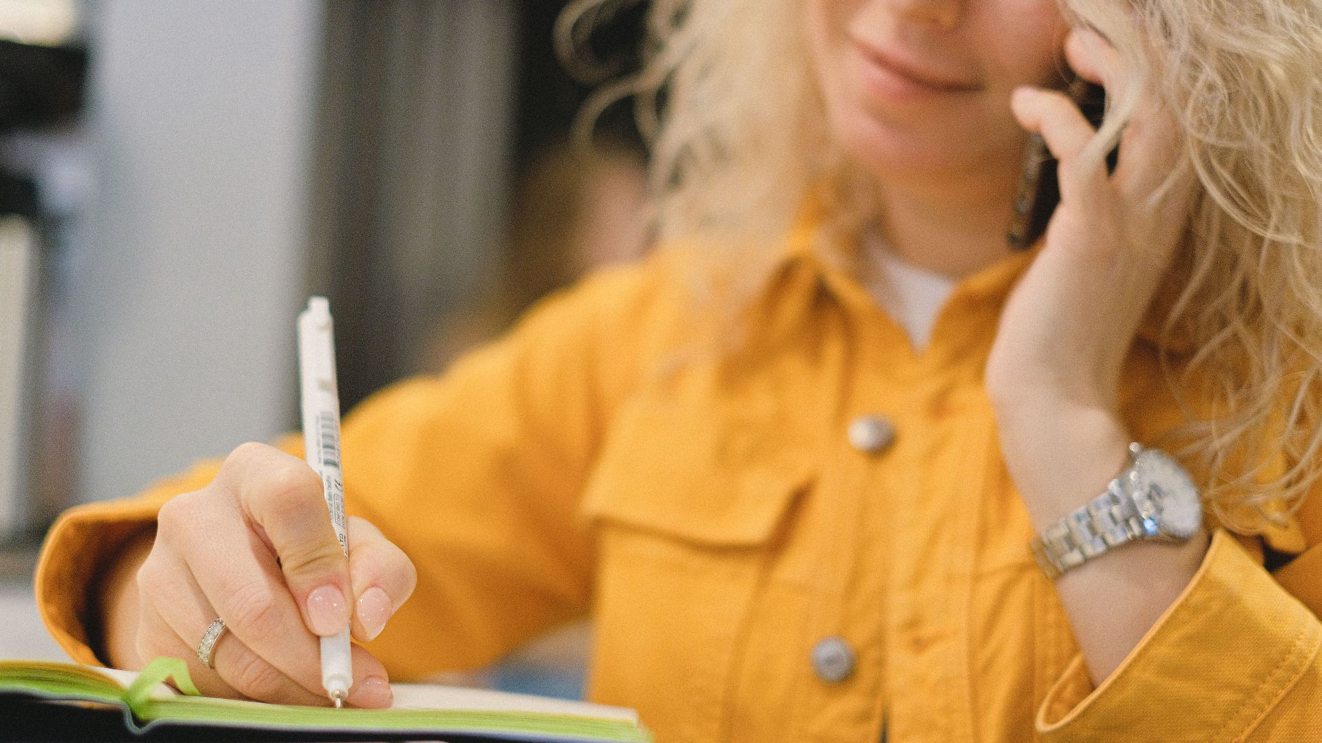 Content blond female employee writing notes while talking on cellphone on blurred background