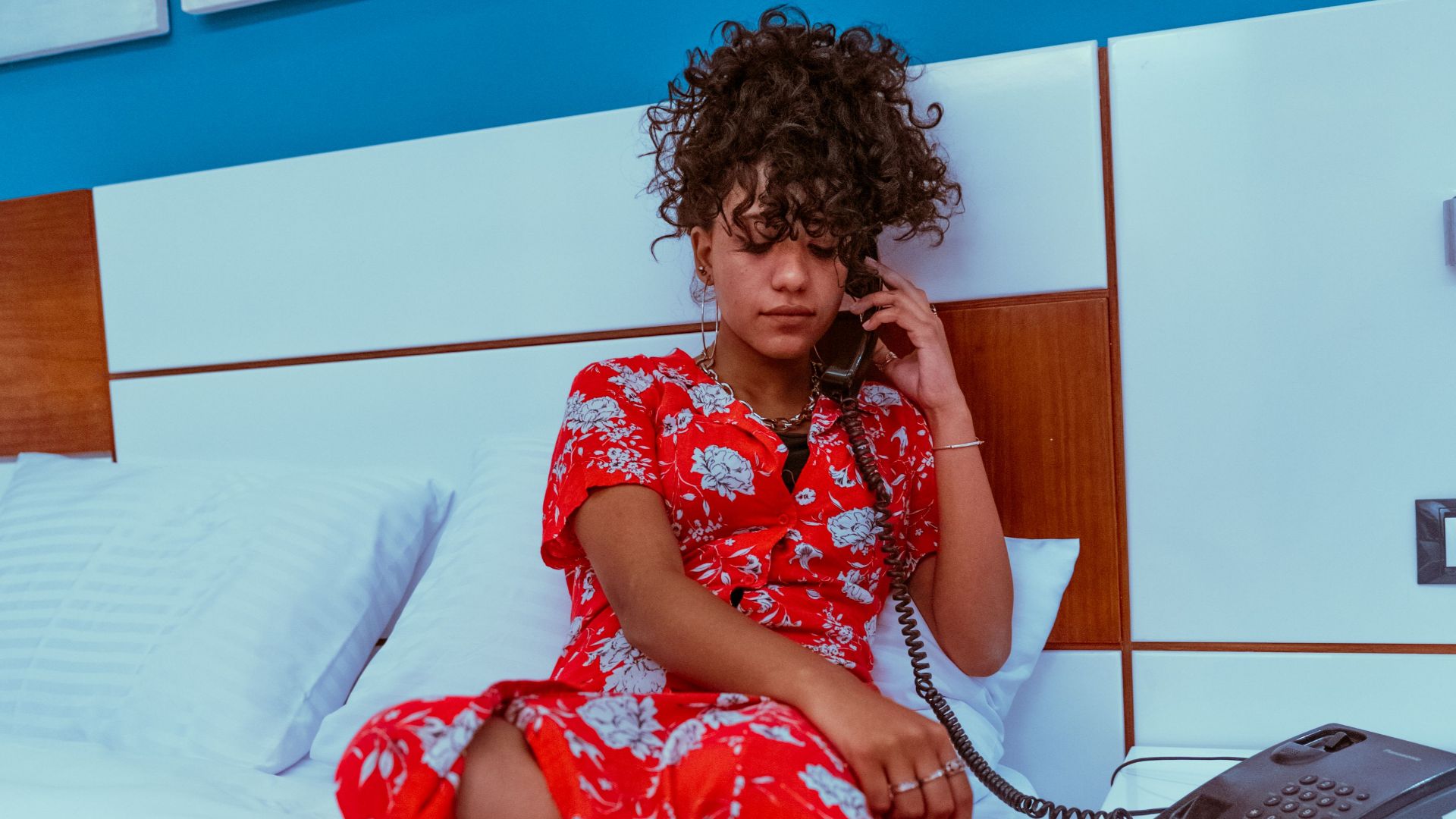 Young woman in vibrant red dress talks on hotel phone while sitting on bed.