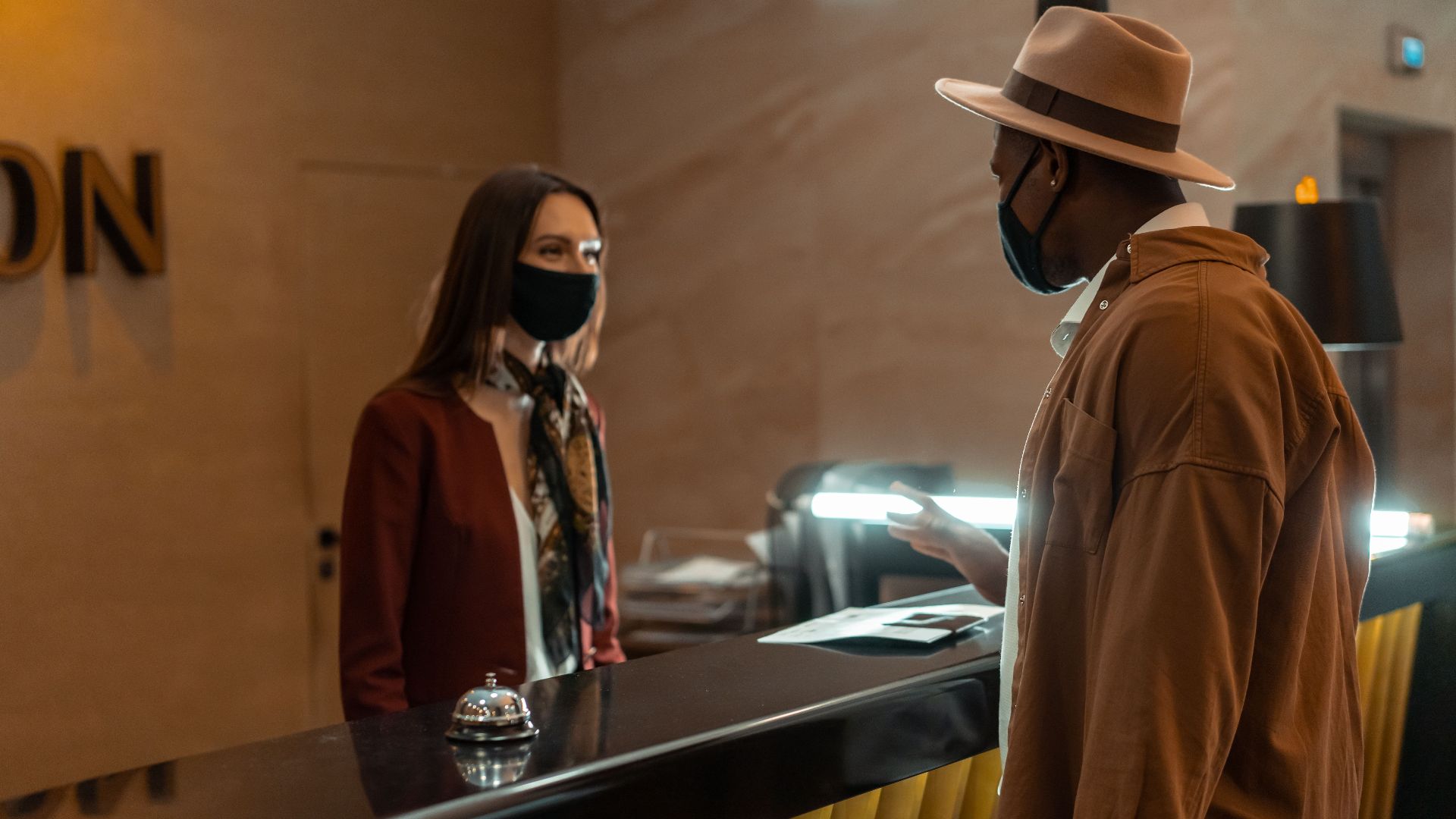 A man at a hotel reception talking to a staff member. Both wear face masks.