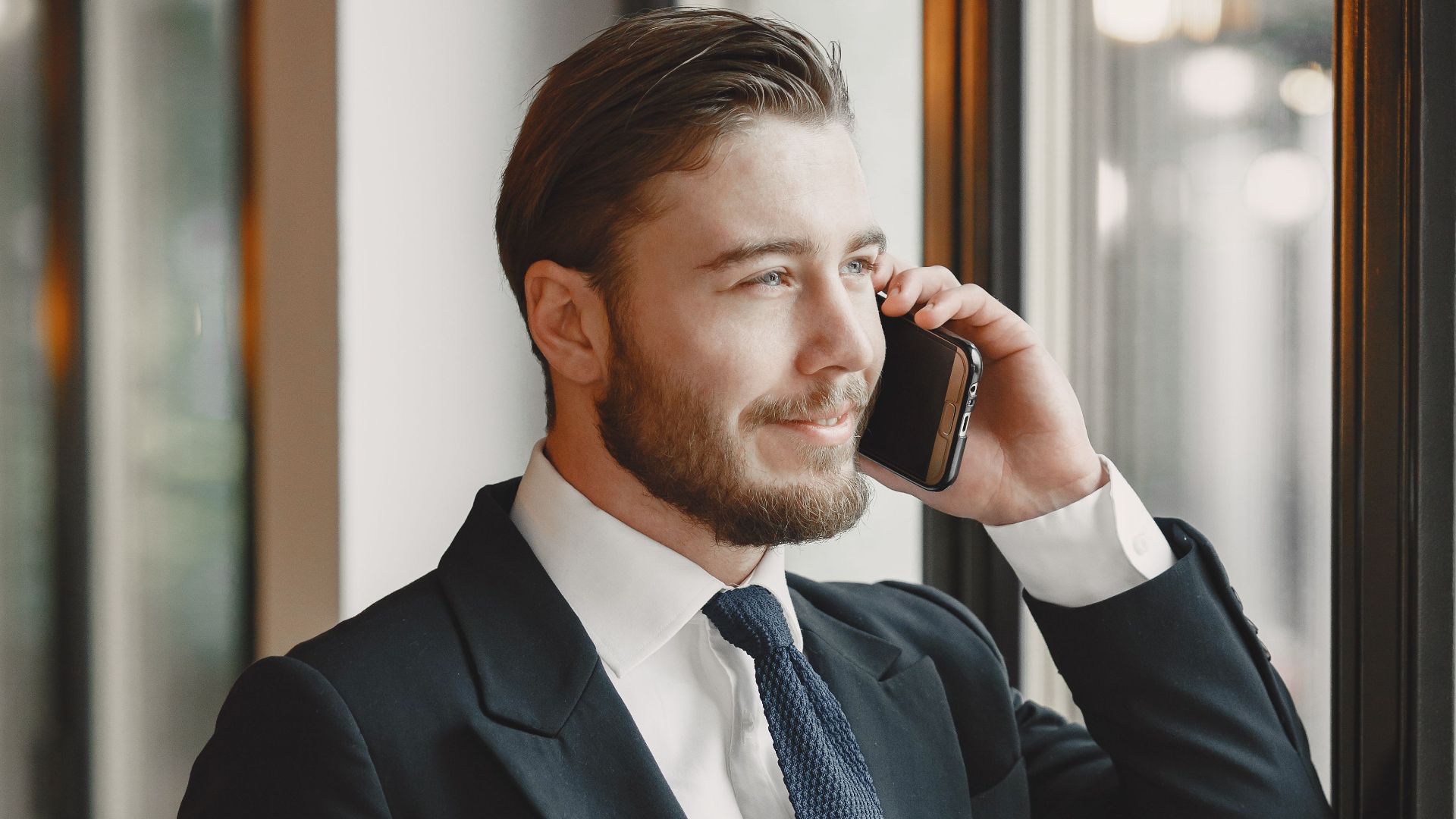 A businessman in a suit talks on the phone by a window, exuding confidence and professionalism.