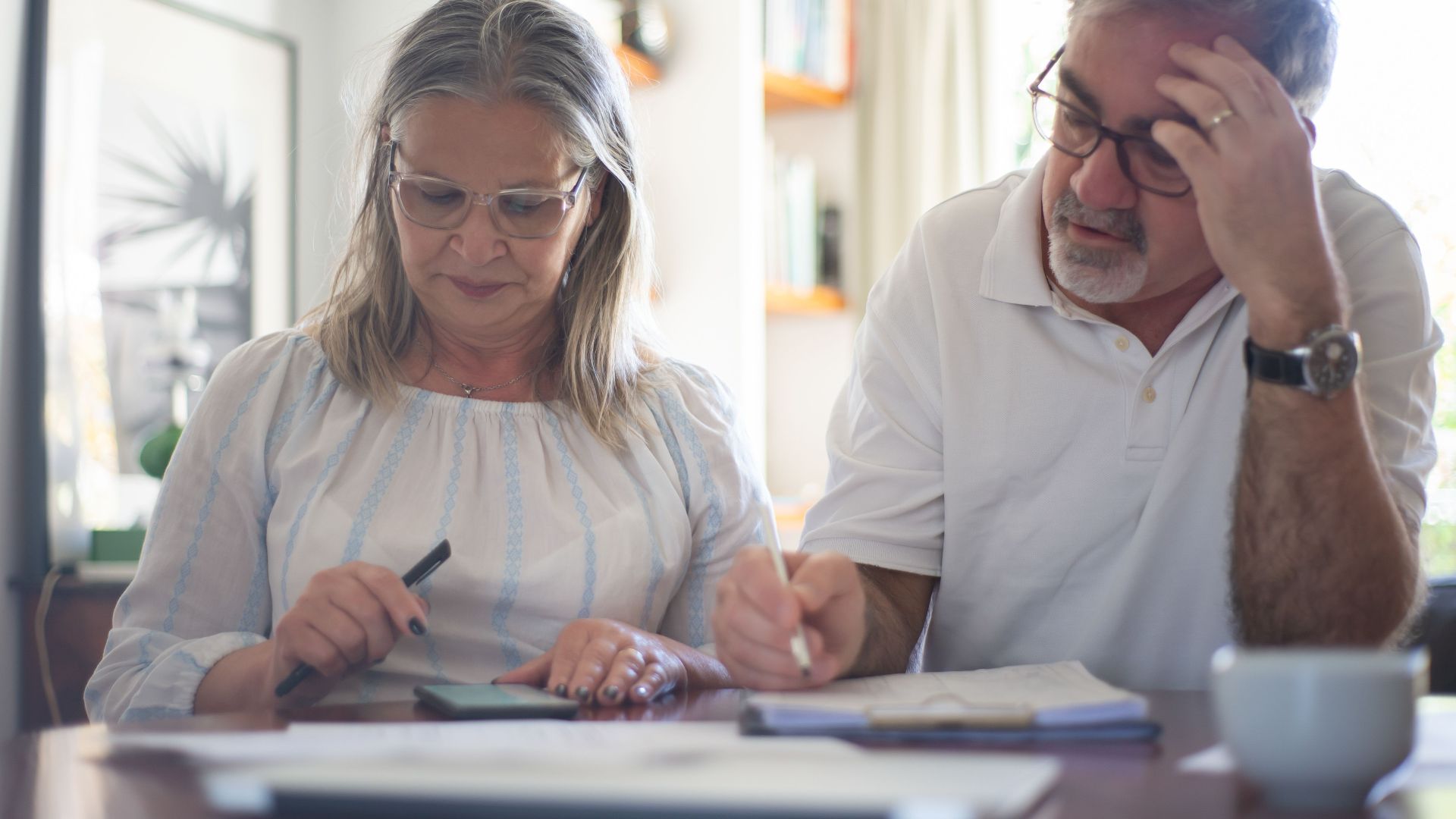 Senior couple calculating expenses at home office desk with documents and notes.
