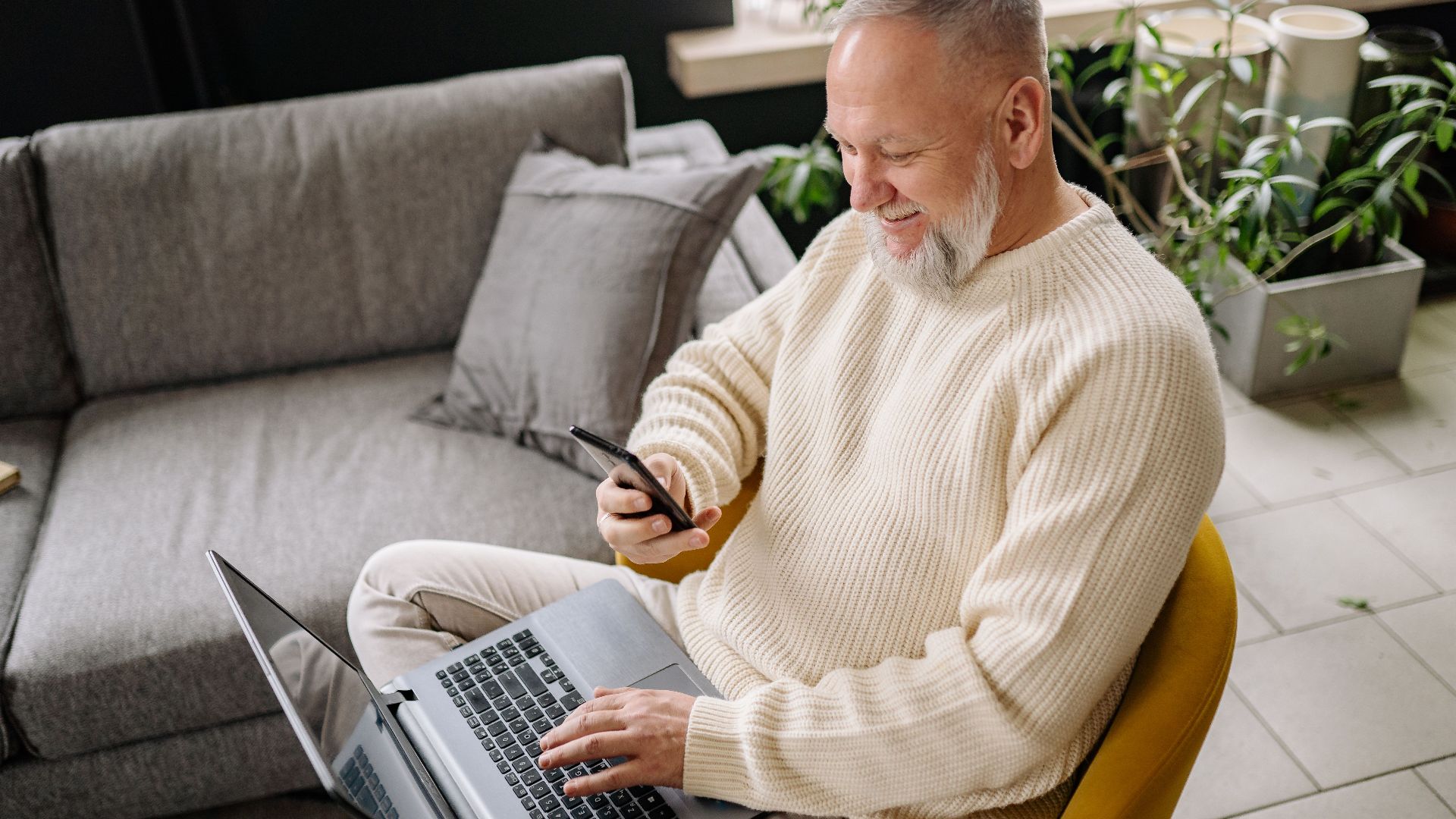 A smiling elderly man sits comfortably indoors, using both a phone and laptop, with a cozy ambiance.