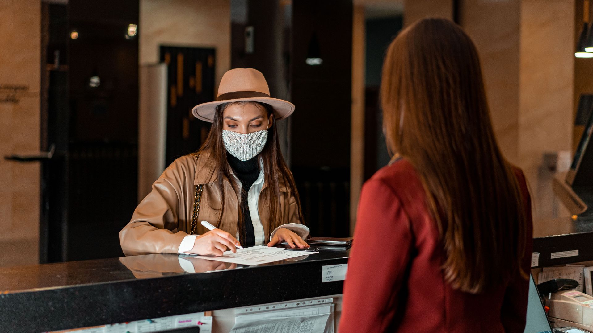 A woman in a hat and face mask checks in at a hotel reception, interacting with a receptionist.