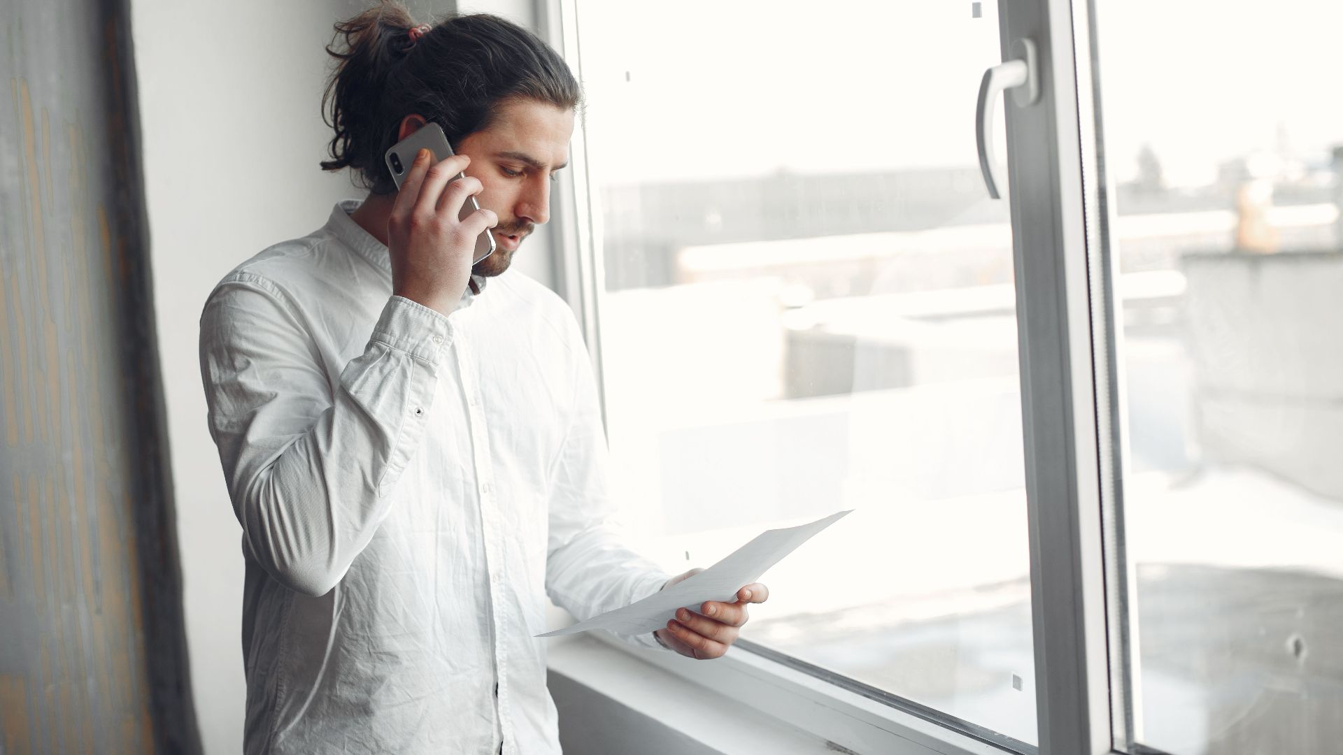 Young man in white shirt, on phone call holding a document, standing by a large window.