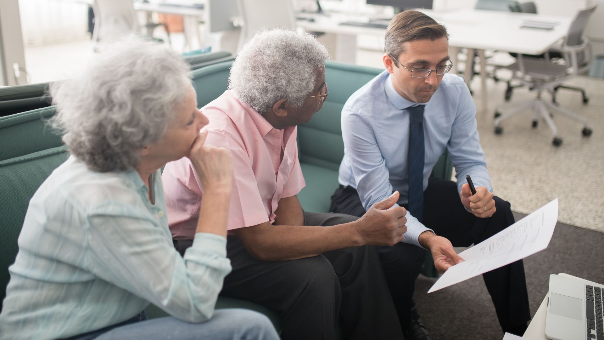 Business professional consults elderly clients in an office setting. Collaborative discussion, paperwork visible.