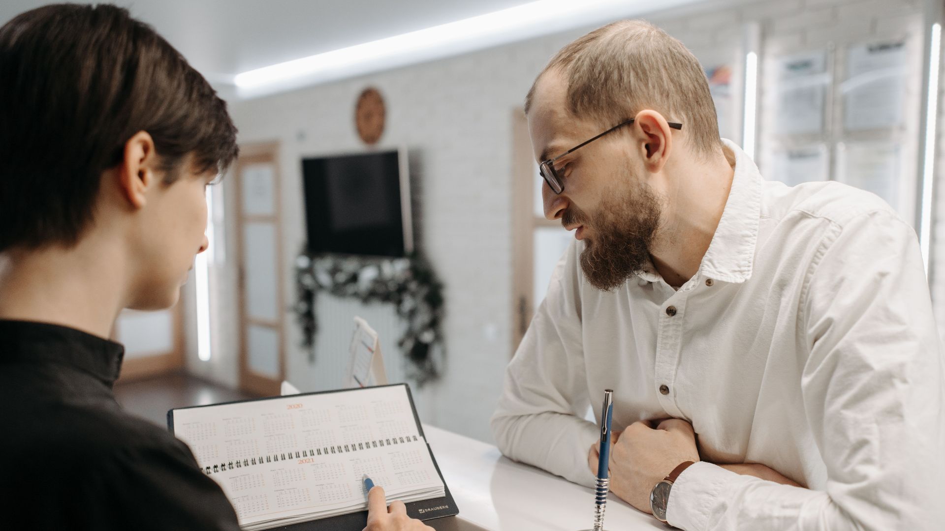 A receptionist and client converse over an appointment book at a clinic reception desk.