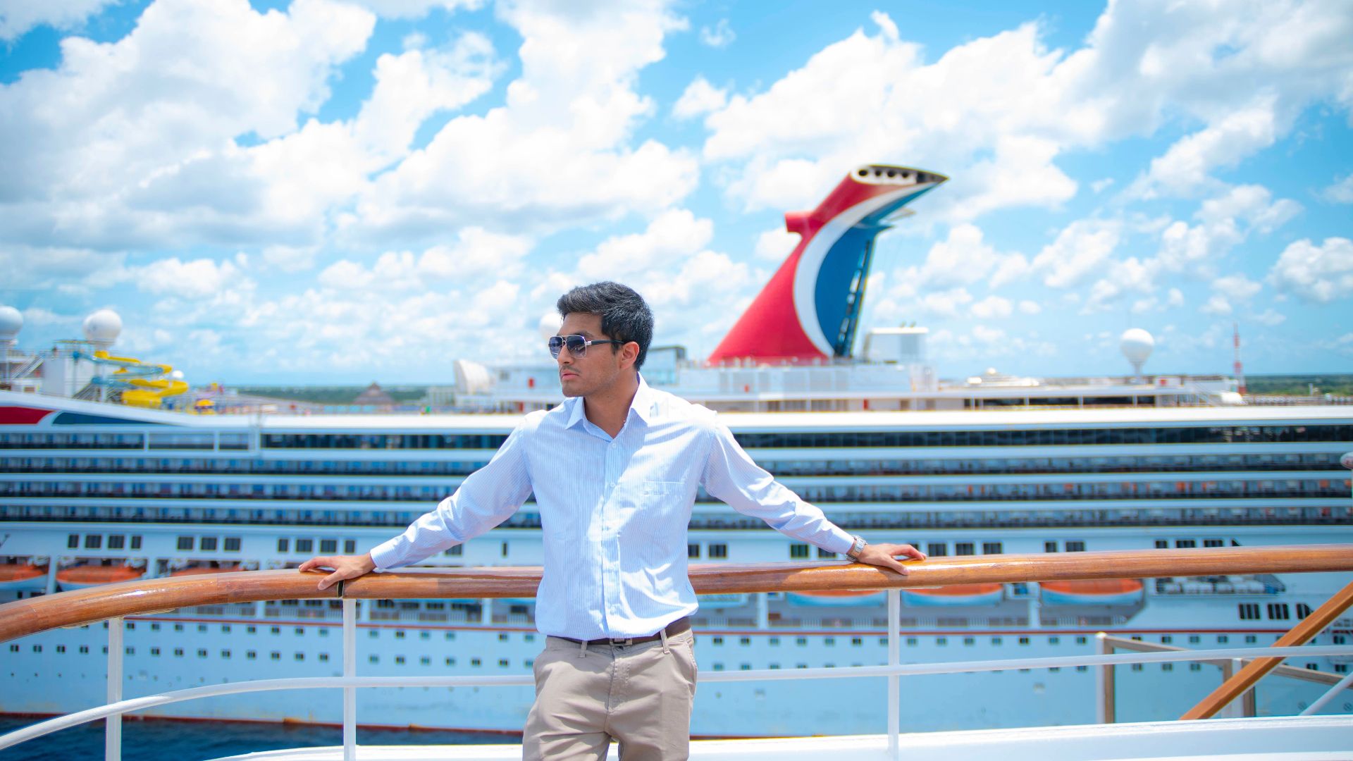 A man in sunglasses and white shirt enjoys a sunny day on a cruise ship deck with puffy clouds overhead.