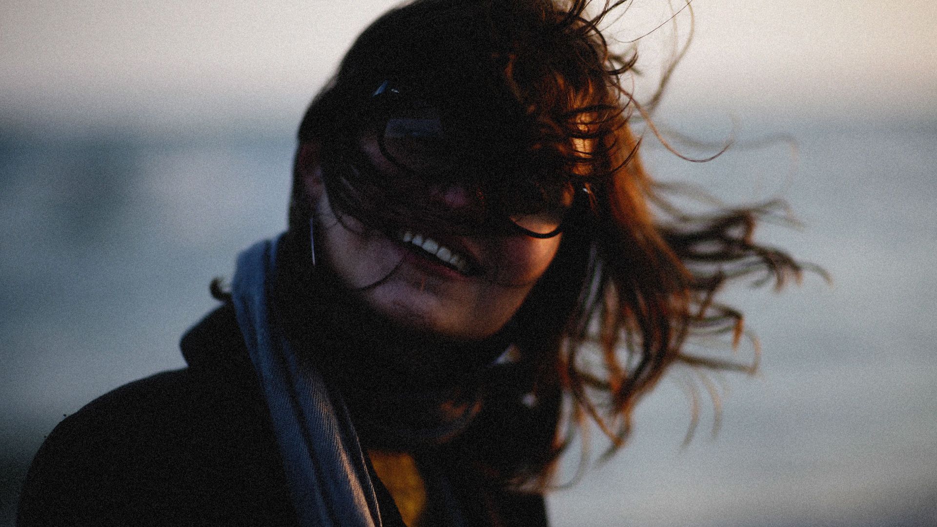A woman with flowing hair and glasses smiles in a windy outdoor setting.