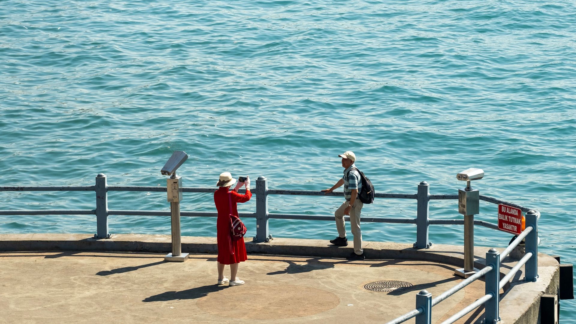 A woman photographs a man on a sunny pier by the sea.