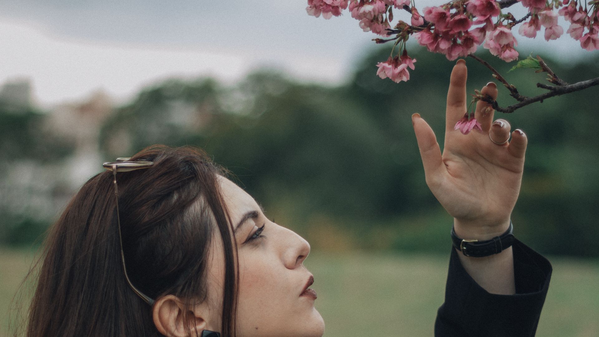 Woman in black outfit reaching for pink cherry blossoms outdoors.