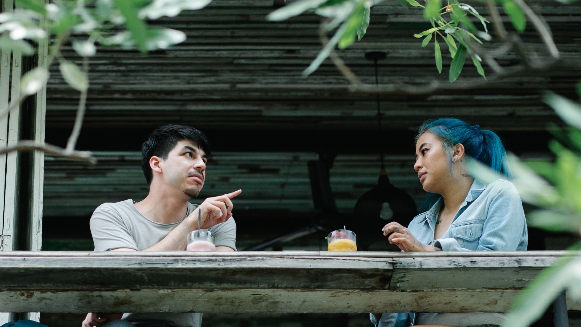 A couple enjoys drinks and conversation at an outdoor café table surrounded by greenery.