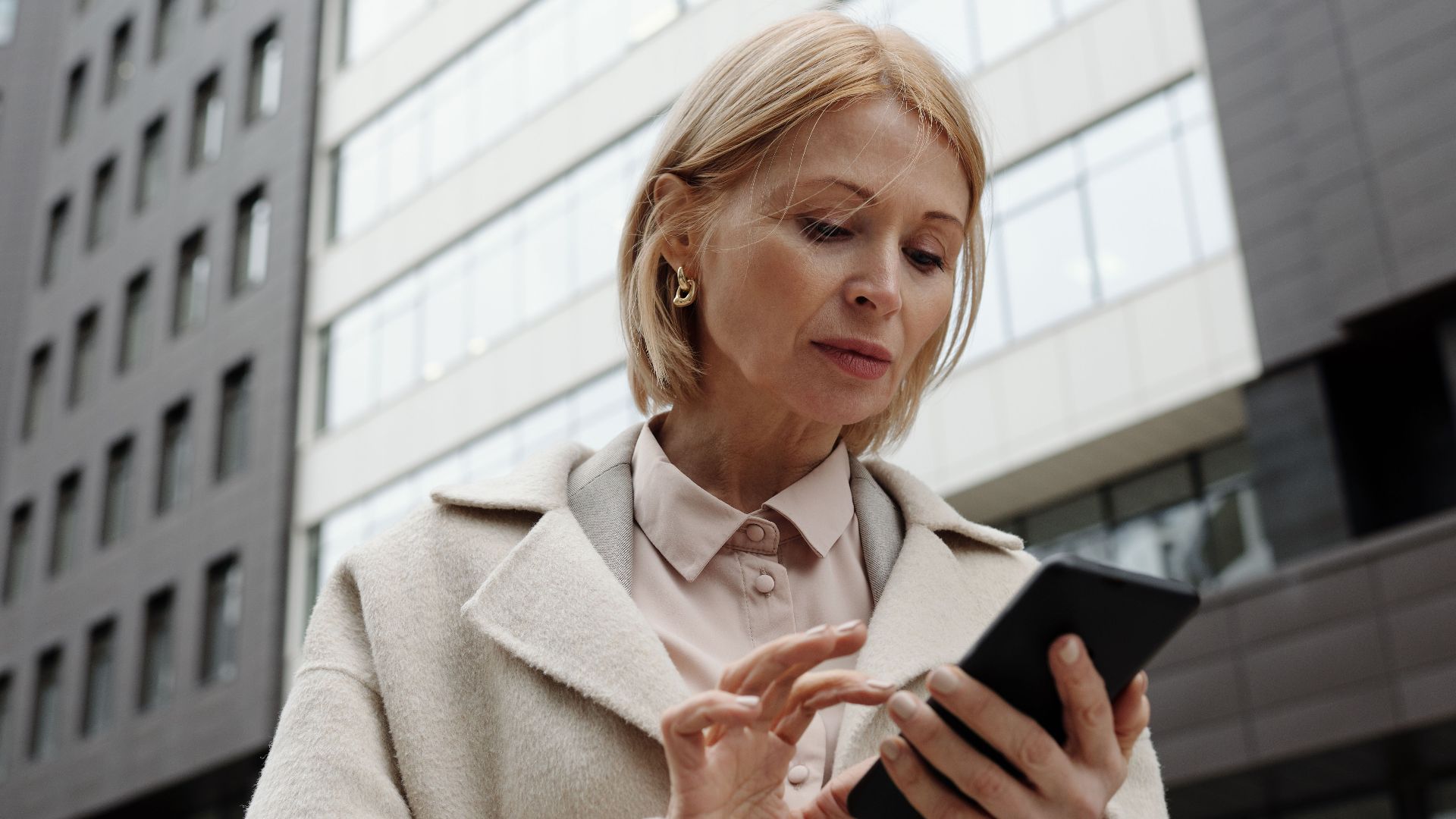 Woman in coat using smartphone in front of modern building. Professional and focused expression.