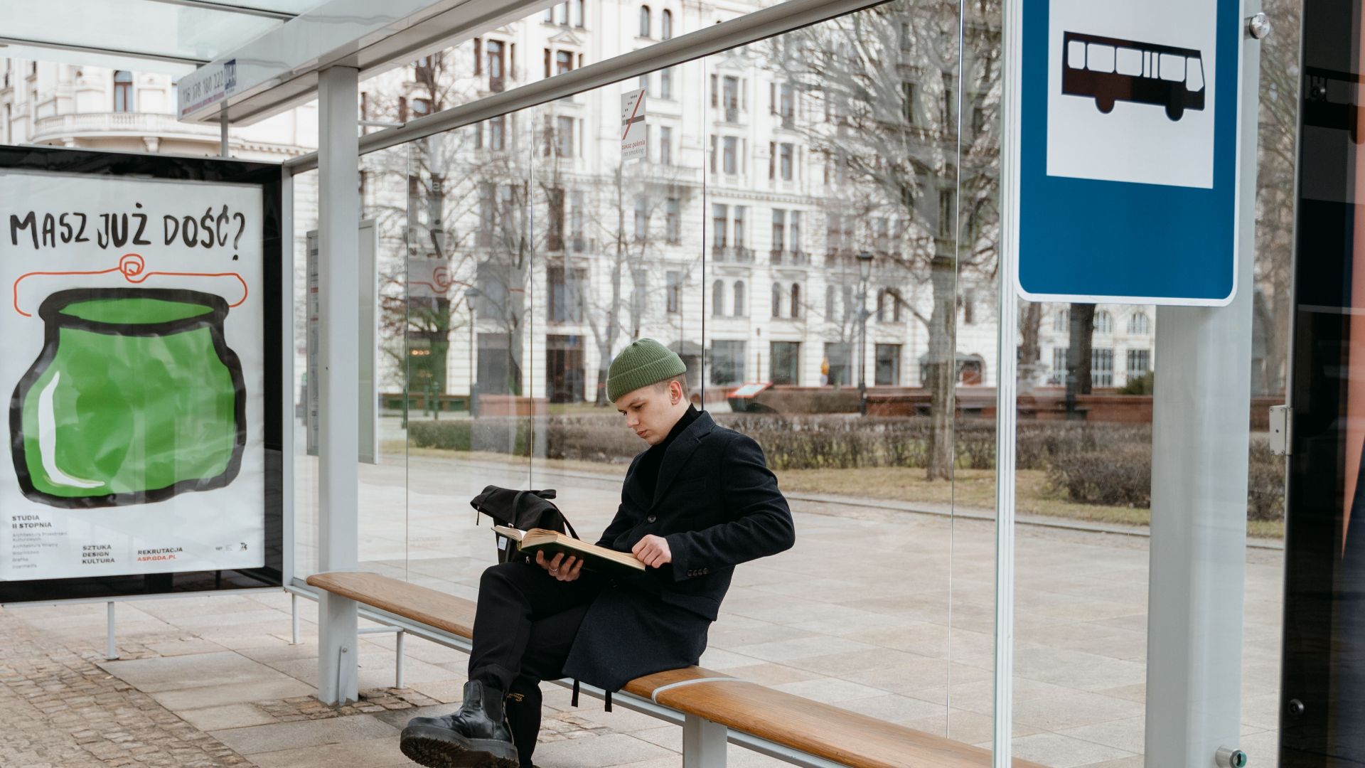 Man in coat reading a book while sitting at an outdoor bus stop on a cobbled street.