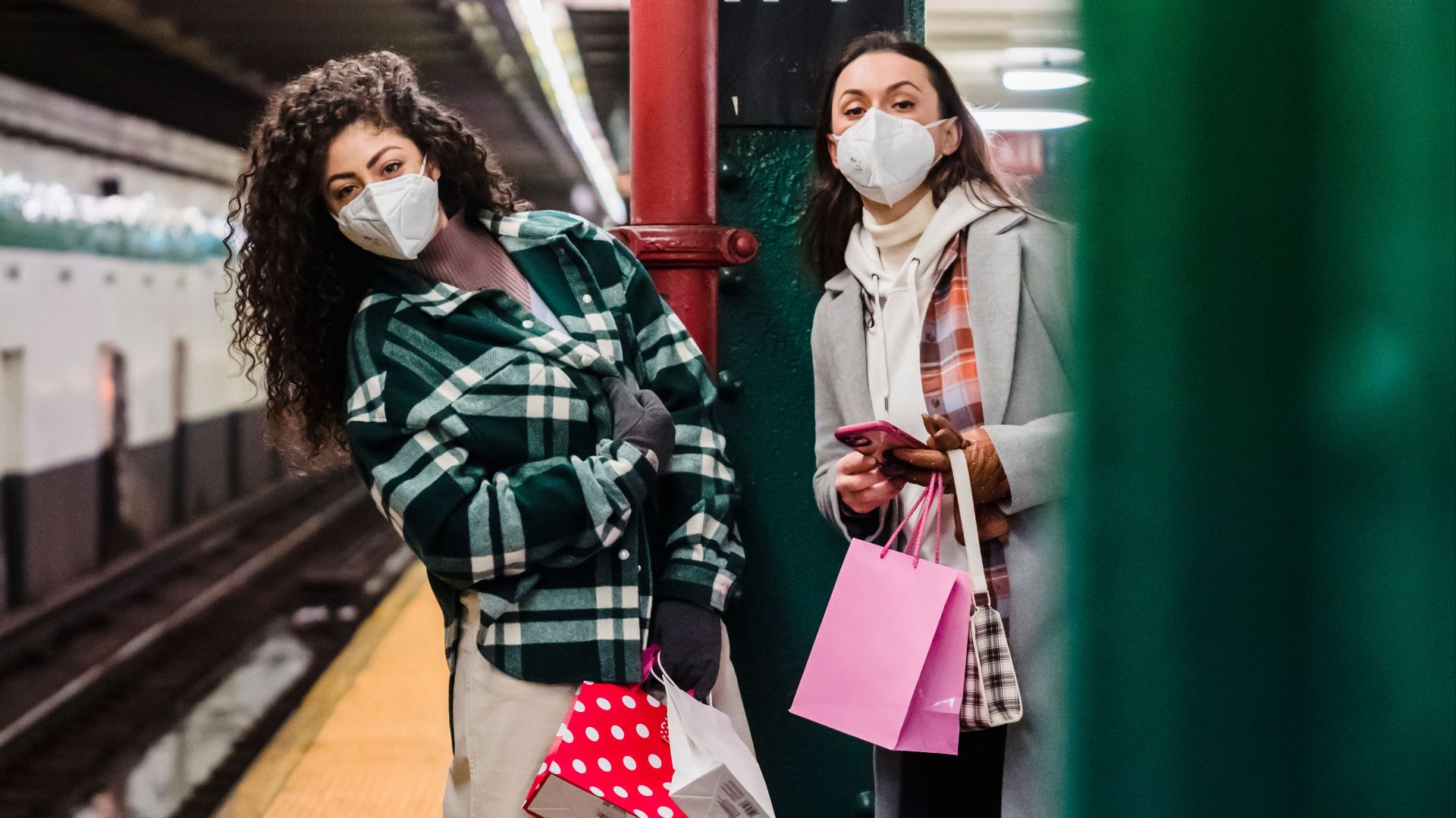 Young female friends in outerwear and protective masks with cellphone and purse with paper bags looking at camera while standing on light metro station