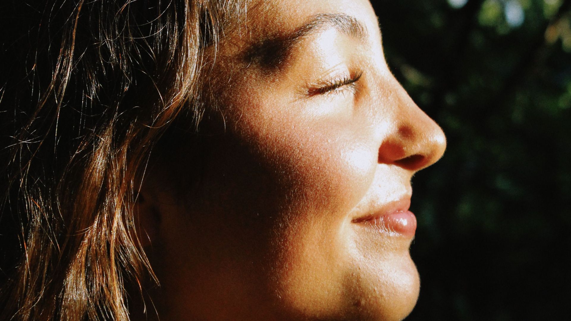 Profile portrait of a young woman smiling in sunlight, embracing nature outdoors.