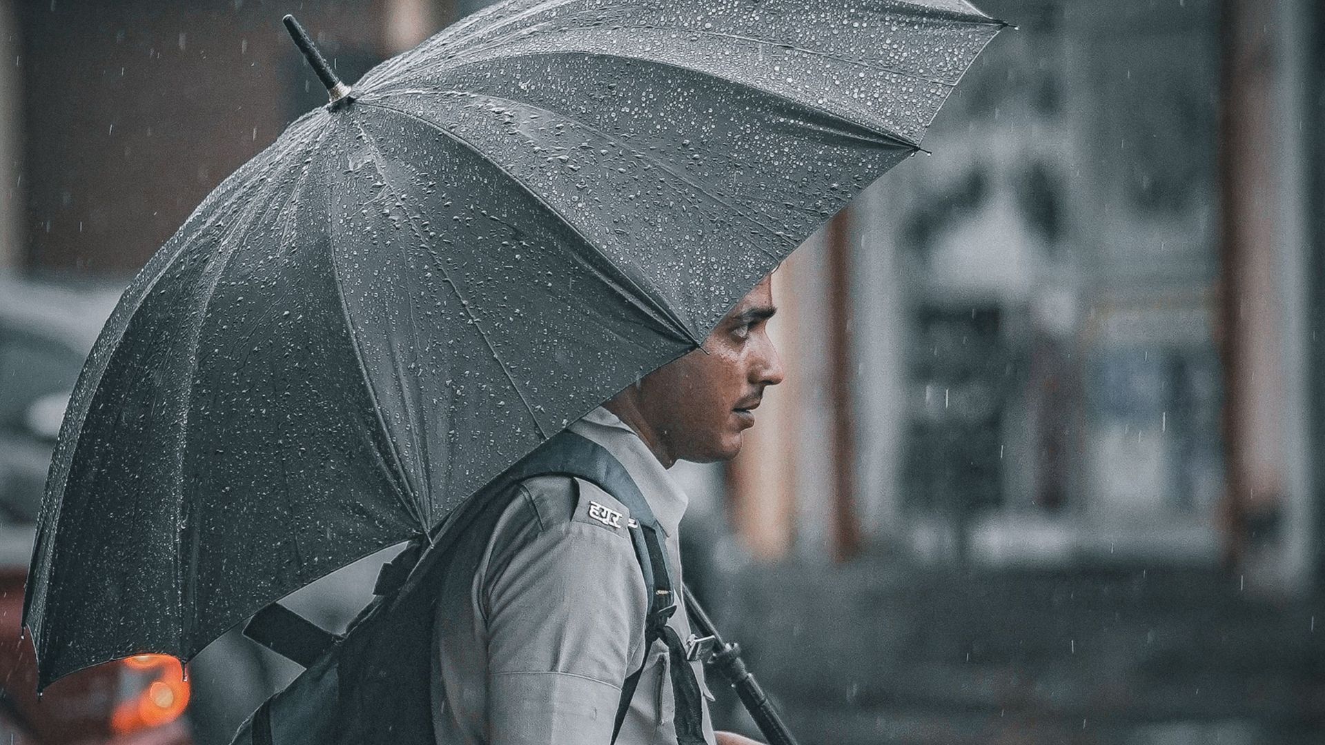 A man wearing a uniform is walking through a rainy street while holding an umbrella and carrying a backpack.