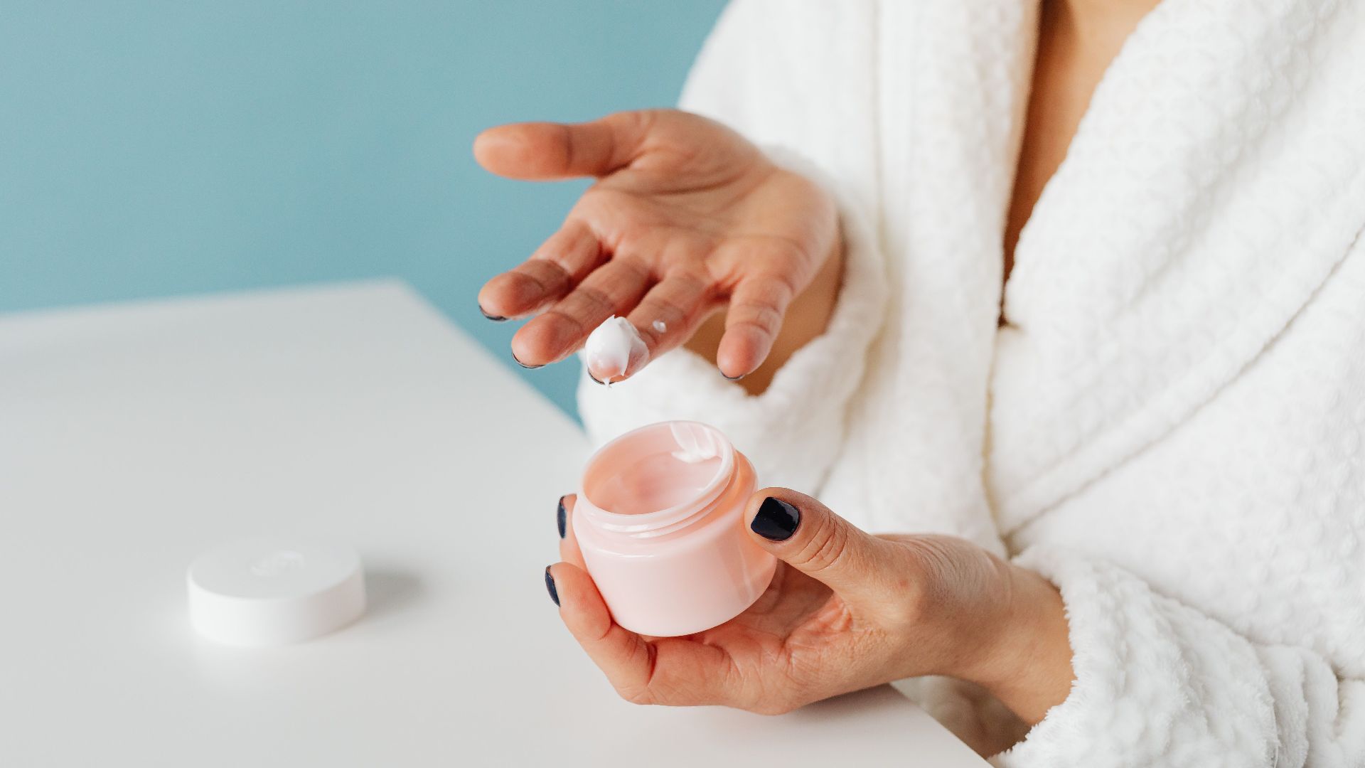 A woman in robe applying skincare cream from an open jar, highlighting moisturizing routine.