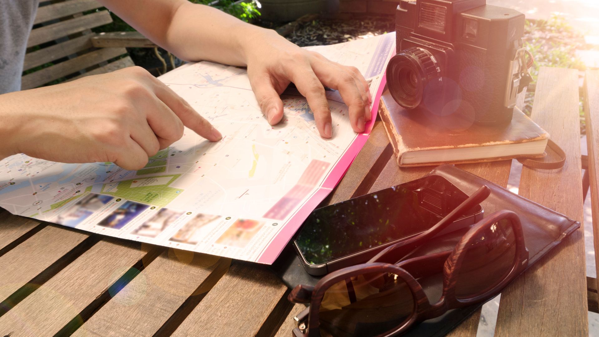 Traveler pointing at map on wooden table with camera and accessories under sunlight.