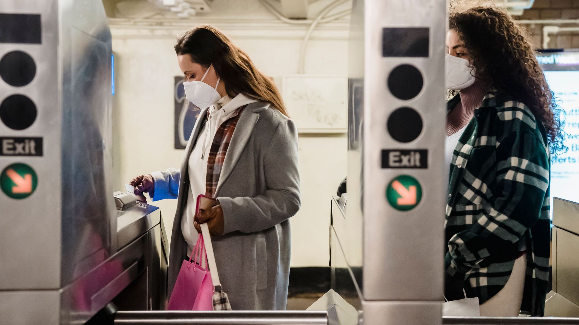 Young women in warm clothes and protective masks with shopping bags near turnstile in bright subway station