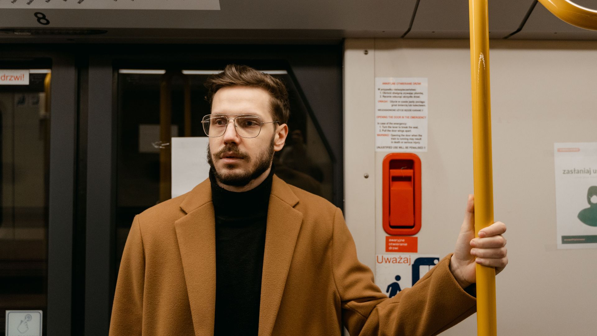 A man wearing a brown coat and glasses stands inside a subway train, holding a handrail.