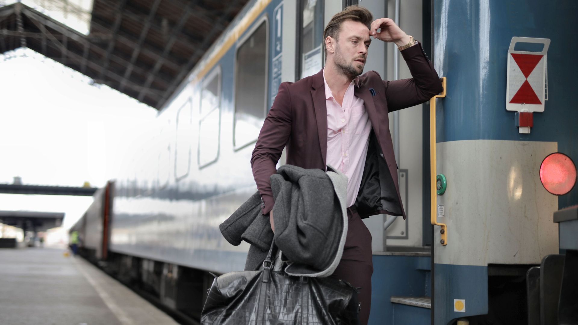 A man in a suit, carrying bags, looks stressed as he boards a train at a railway station.